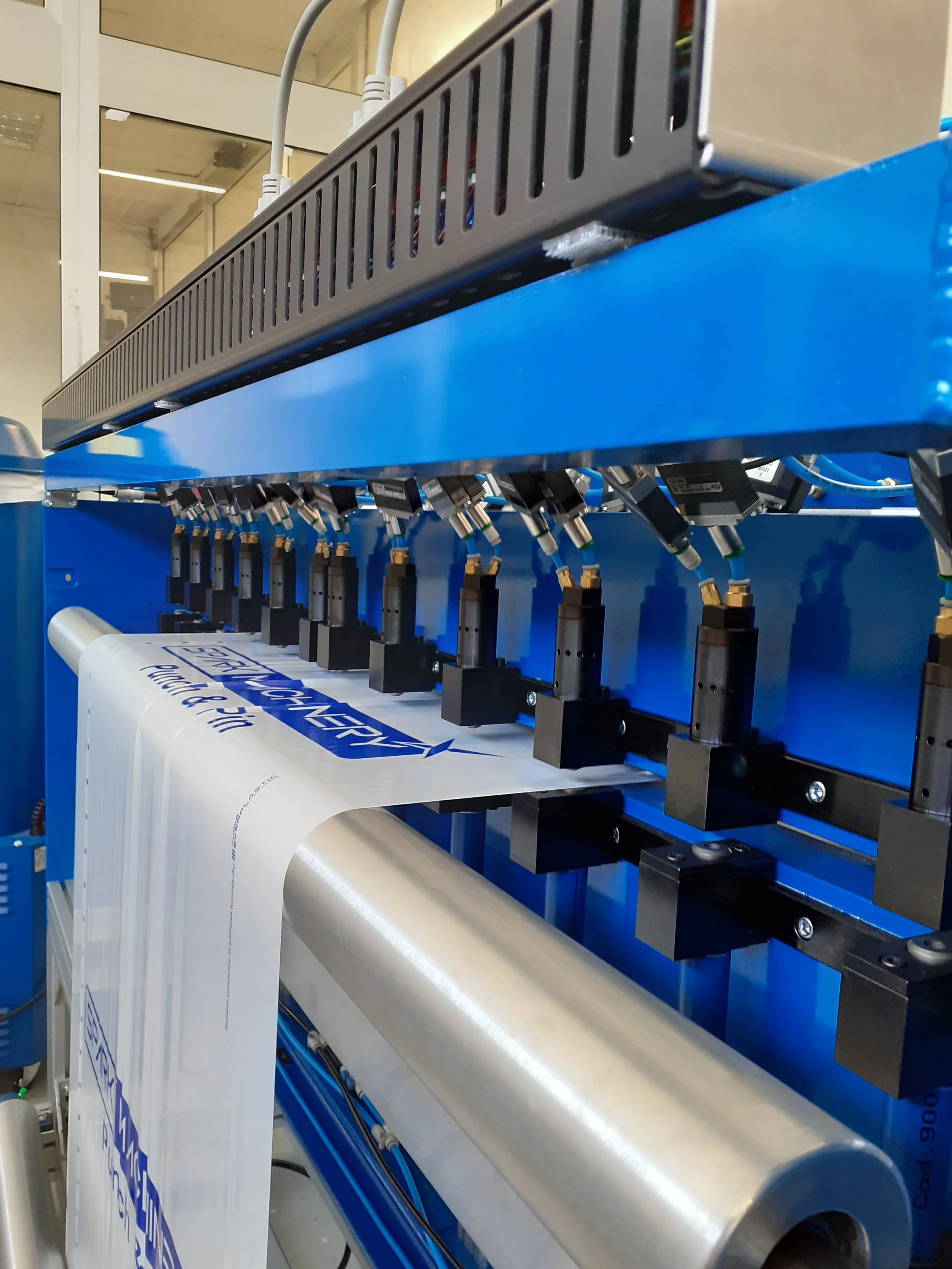 Industrial machine with multiple black components connected to a roll of material, with a white printed label partially visible.