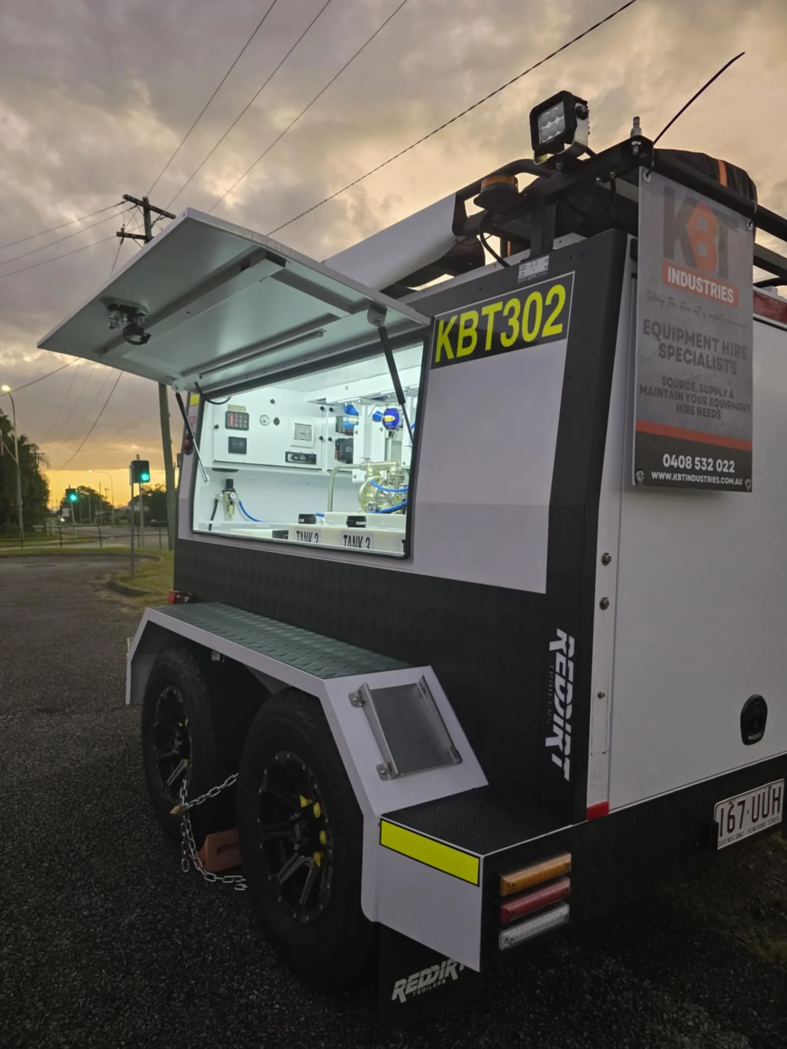 Mobile equipment rental trailer with open service window, tools, tanks, and equipment inside, parked on a street during sunset with utility power lines and traffic lights in the background.