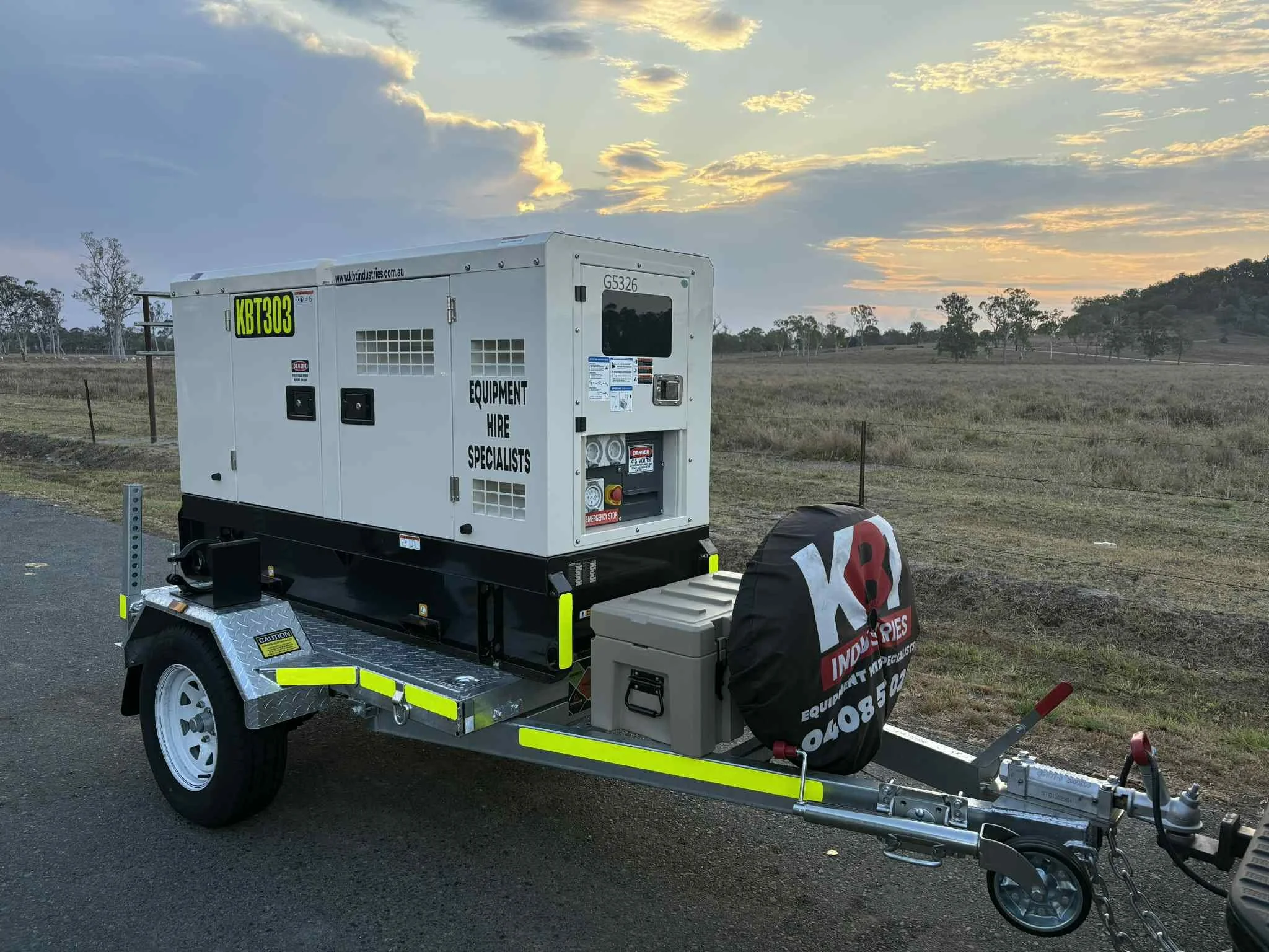 A portable generator on a trailer parked on an open field at sunset. The generator has signs indicating equipment hire services.