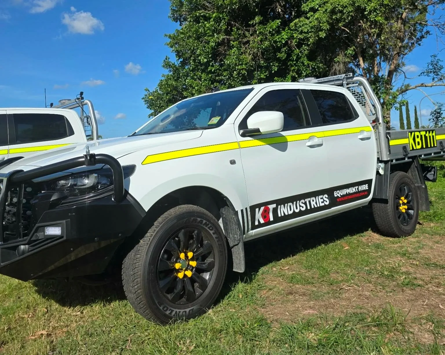 White utility truck with black and yellow accents, parked on grass with trees and blue sky in the background. The truck has a logo that reads 'KBT INDUSTRIES' and additional markings.