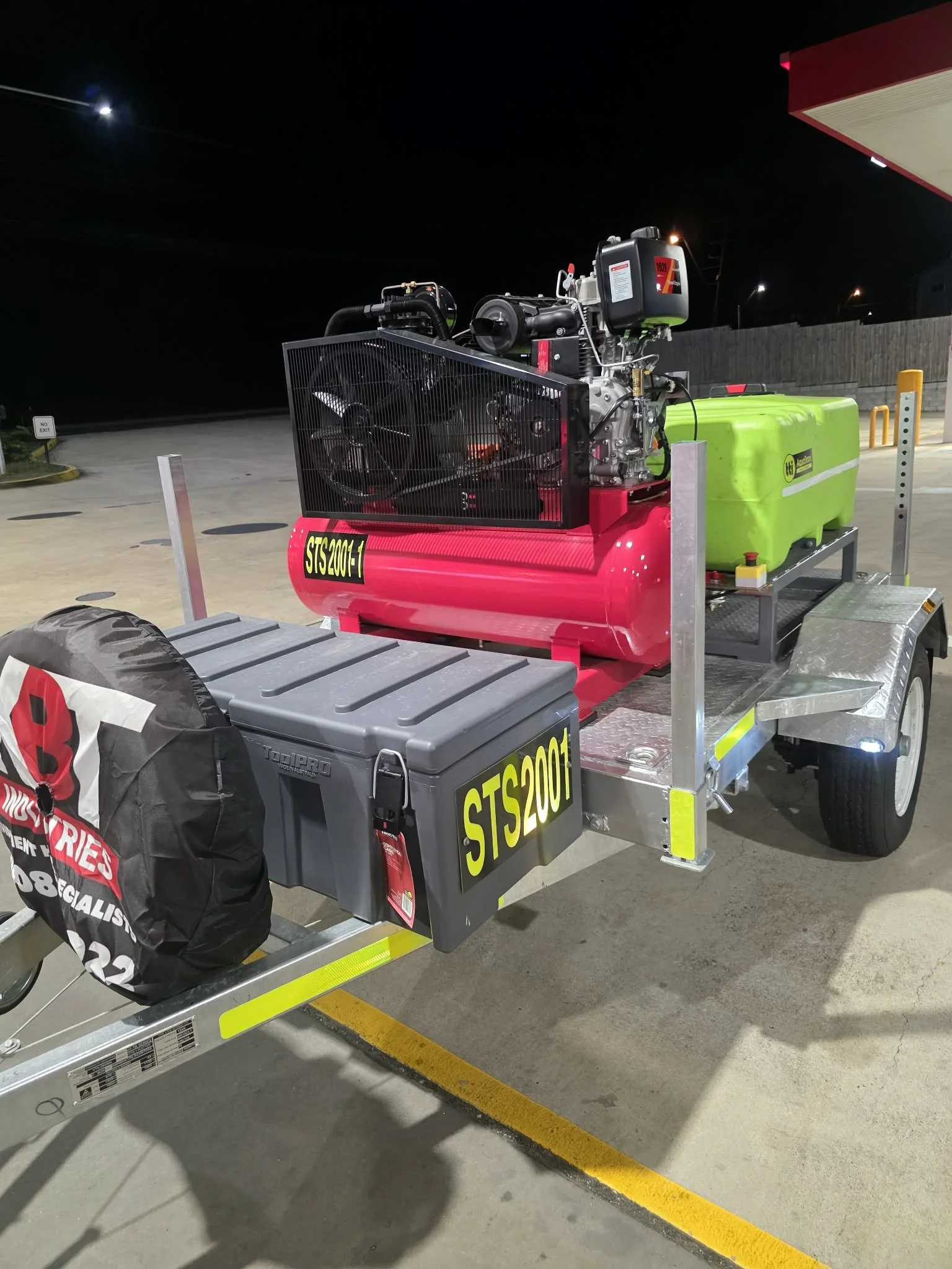 A small trailer carrying a red air compressor, a green water tank, and a large gray toolbox, parked in a lot at night.