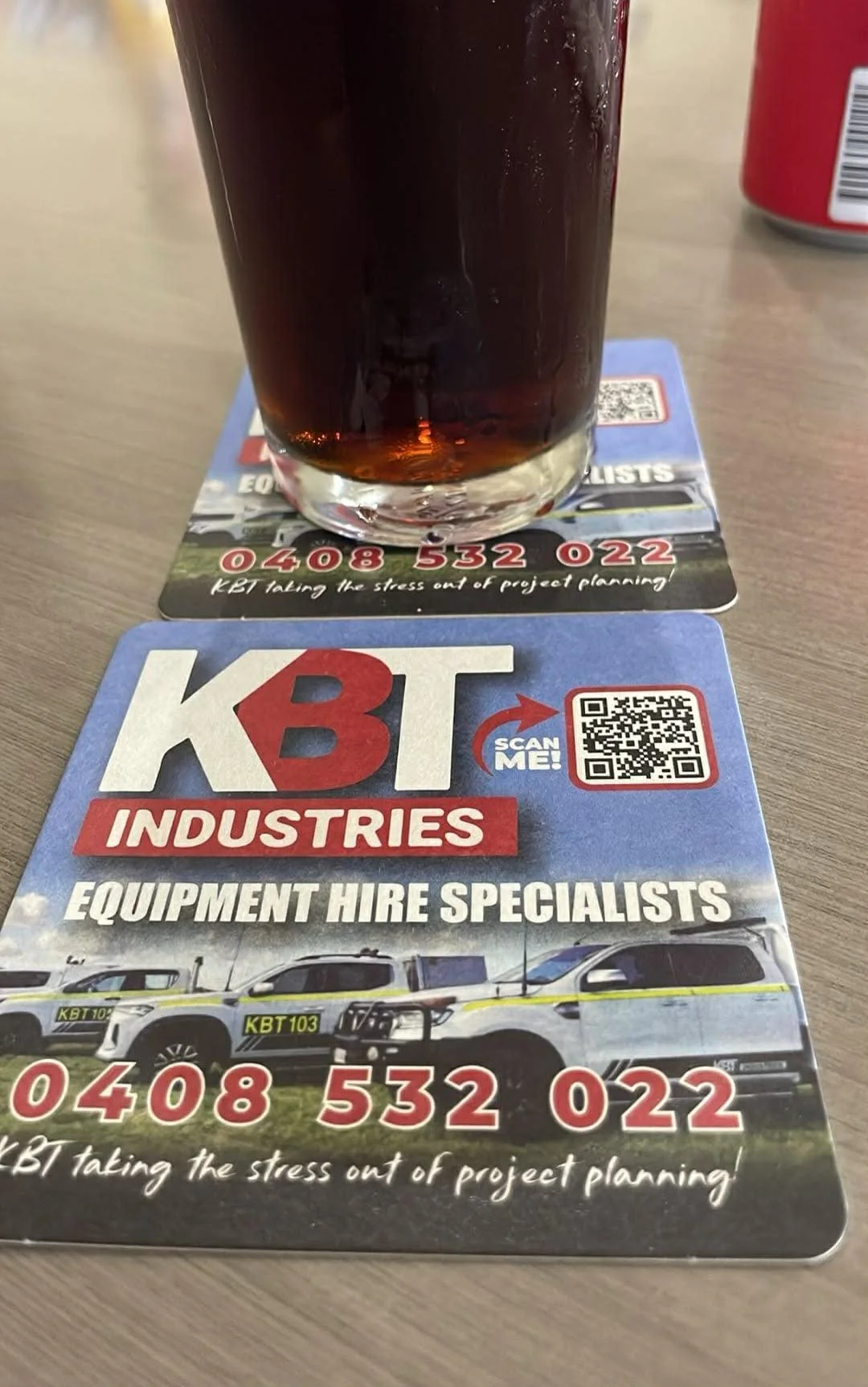 A glass of dark soda on top of two overlapping blue promotional sheets for KBT Industries, featuring trucks, a QR code, and the phone number 0408 532 022.