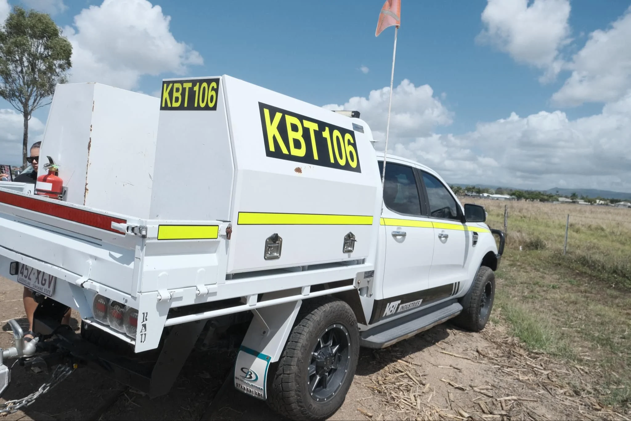 A white utility vehicle with a black and yellow sign on the side and back reading "KBT106." The vehicle is equipped with a red fire extinguisher on the rear and a flag on a pole. It is parked on a dirt area with grass, trees, and a partly cloudy sky in the background.