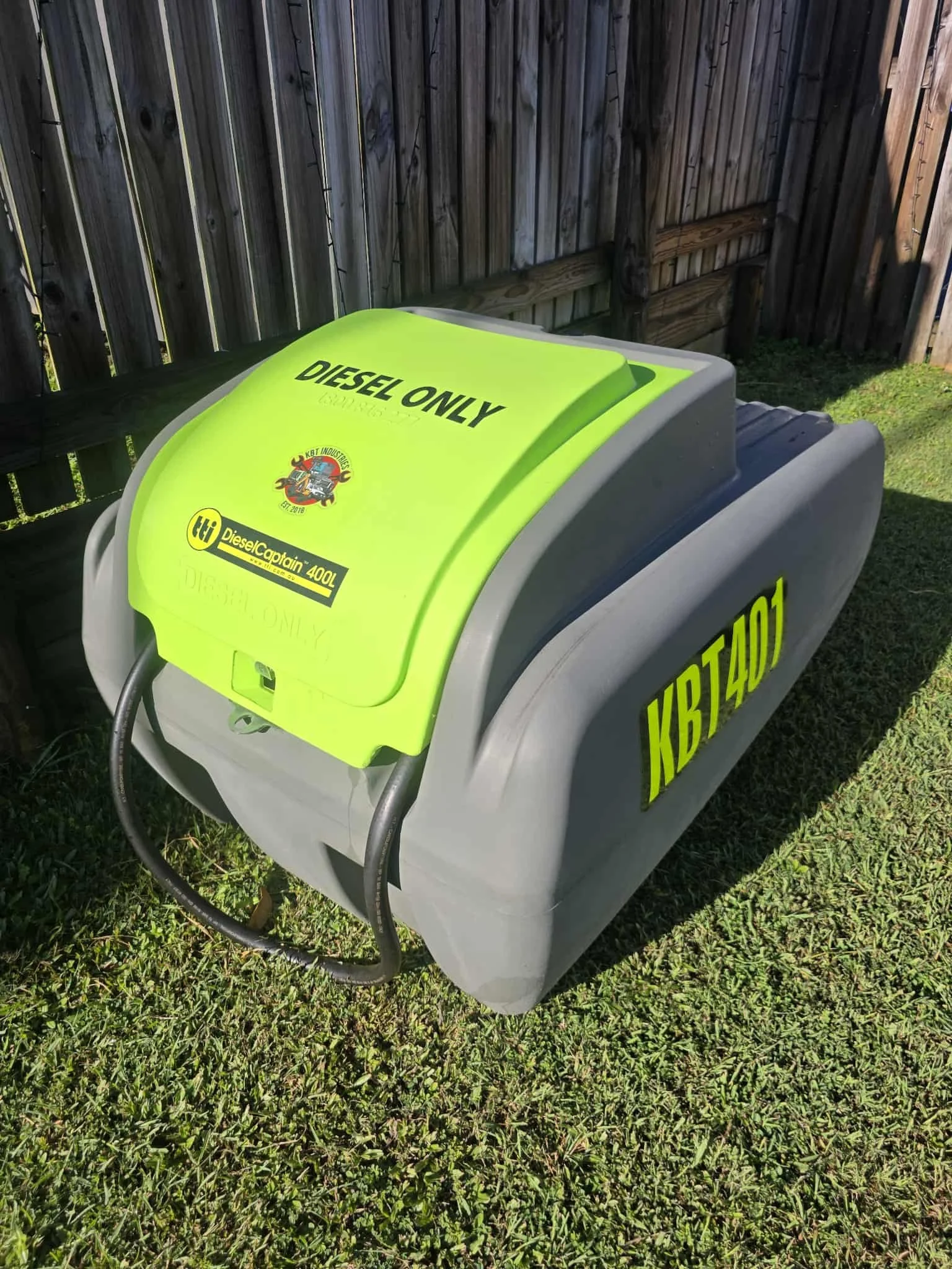 Green and gray diesel fuel storage tank with label 'Diesel Only' and contact information, placed on grass near a wooden fence.