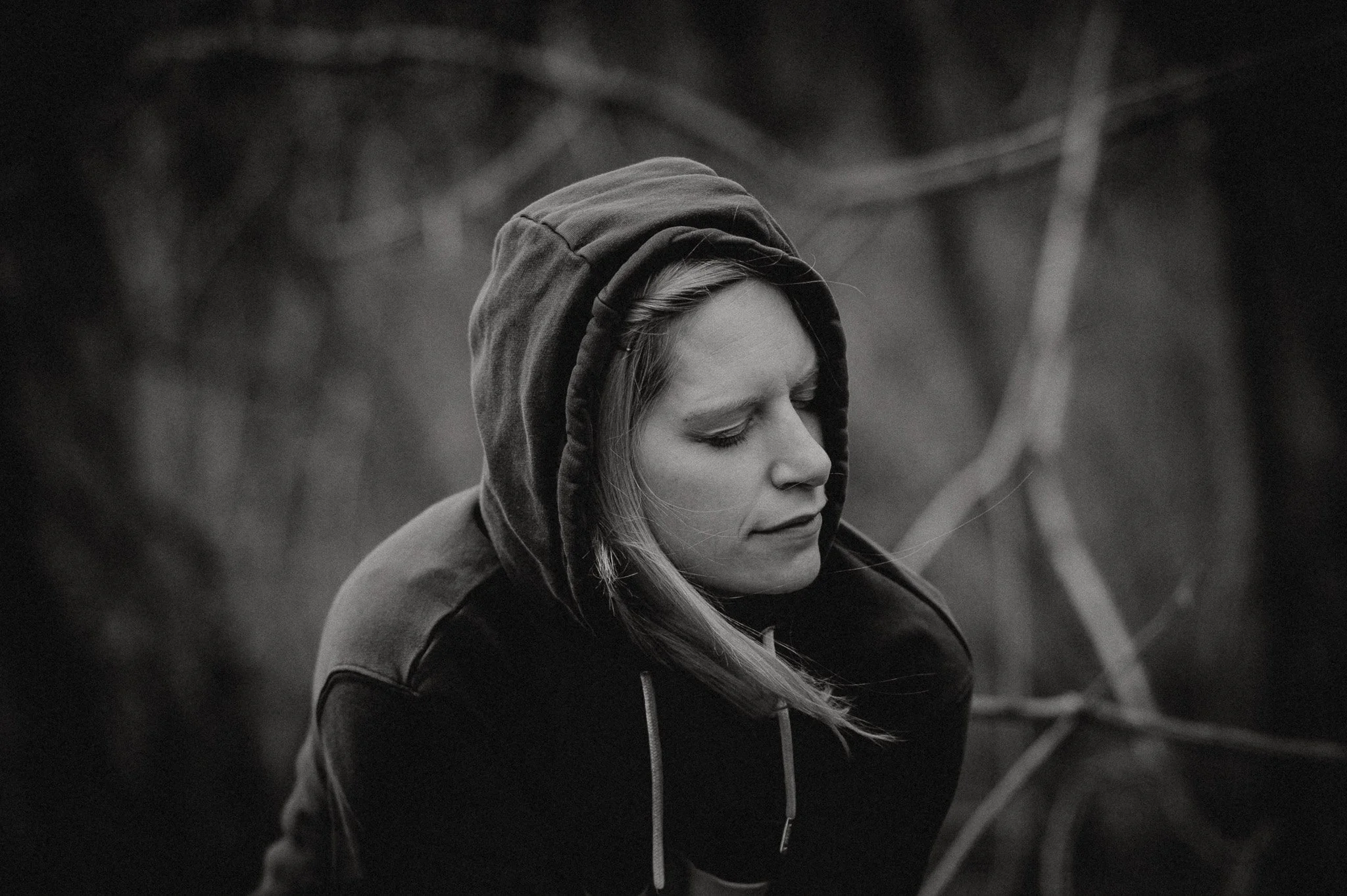 Eine Frau im Hoodie hockt im Wald. Die Kapuze über den Kopf gezogen und die Augen sind geschlossen.