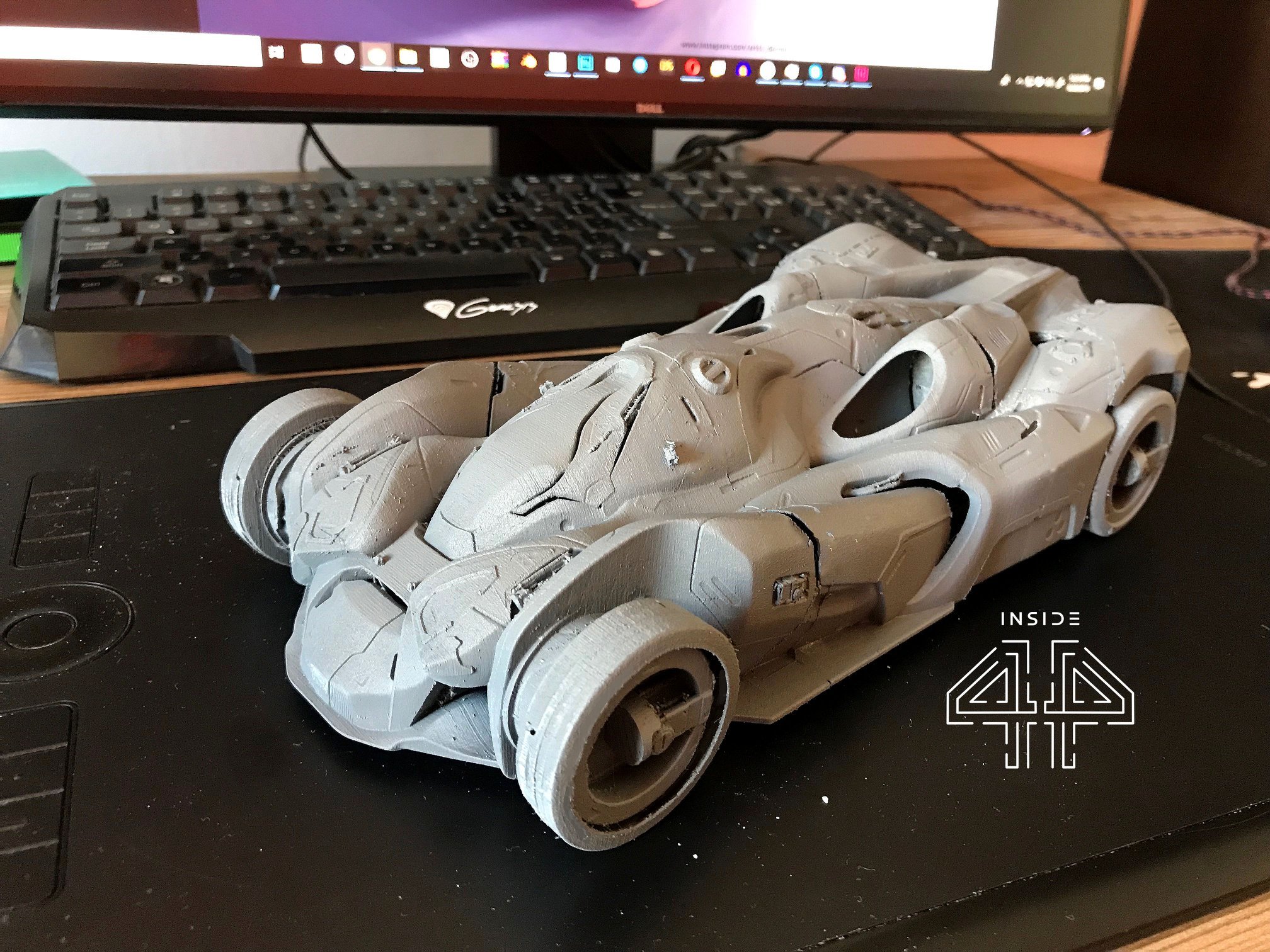 Unfinished gray 3D-printed model of a futuristic car on a black desk mat in front of a computer keyboard and monitor.