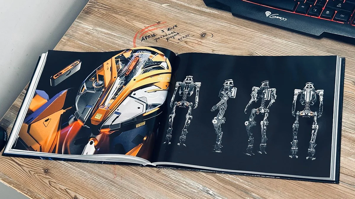 Open book displaying images of futuristic vehicle design on the left page and a robotic skeleton on the right page, placed on a wood desk with a keyboard partially visible in the top right corner.