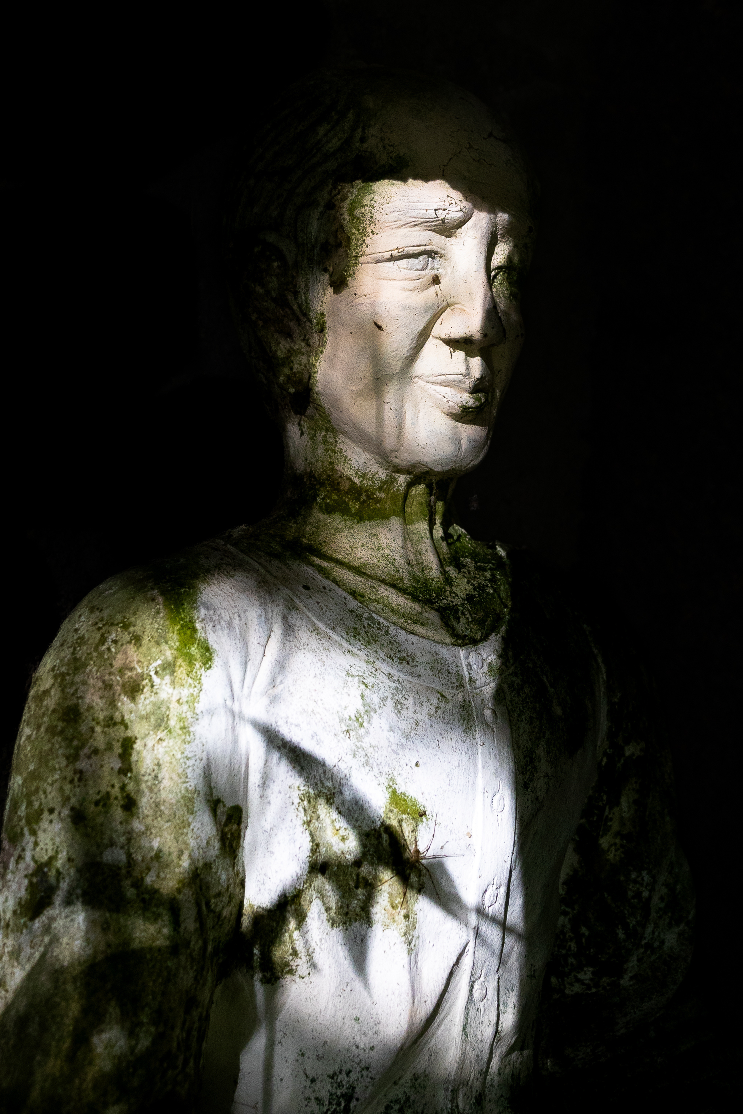 Een oude, mossige houten beeldhouwwerk van een vrouw met gesloten ogen, in een donkere omgeving.