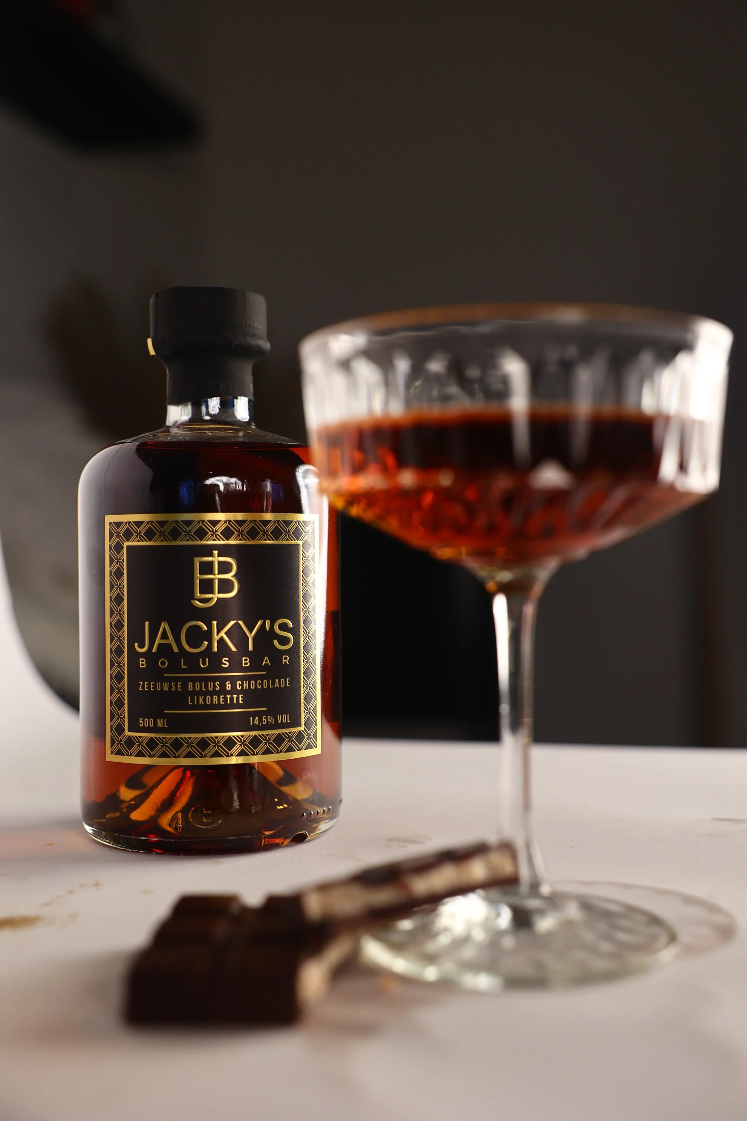 Een flesje Jacky's likeur en een gevuld cocktailglas met een donkere drank, naast stukjes chocolade op een witte tafel.