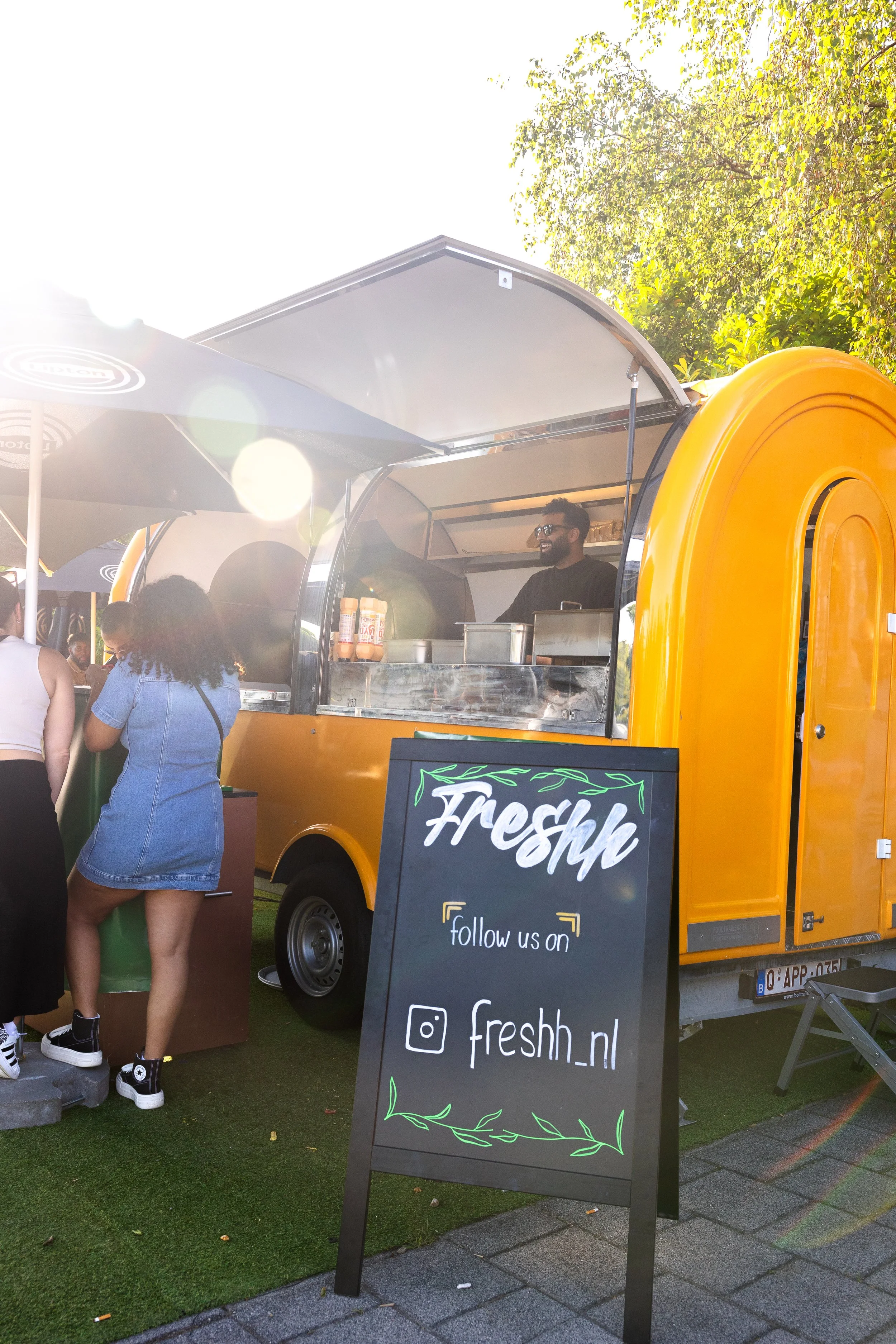 Een foodtruck met een oranje kleur en een man achter het raampje die serveert. Een bord met de tekst 'Freshh' en de instructie 'follow us on' met Instagram-link 'freshh.nl'. Mensen staan in de rij bij de foodtruck. Het is een zonnige dag met groene bomen op de achtergrond.