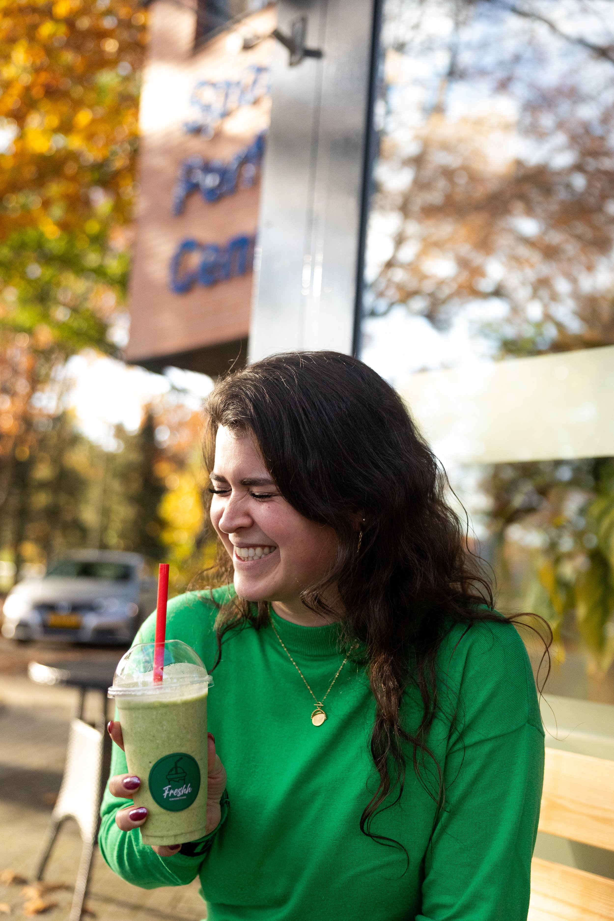 Vrouw in een groene trui die een groene smoothie vasthoudt en lachend zit op een bank buiten in de herfst.
