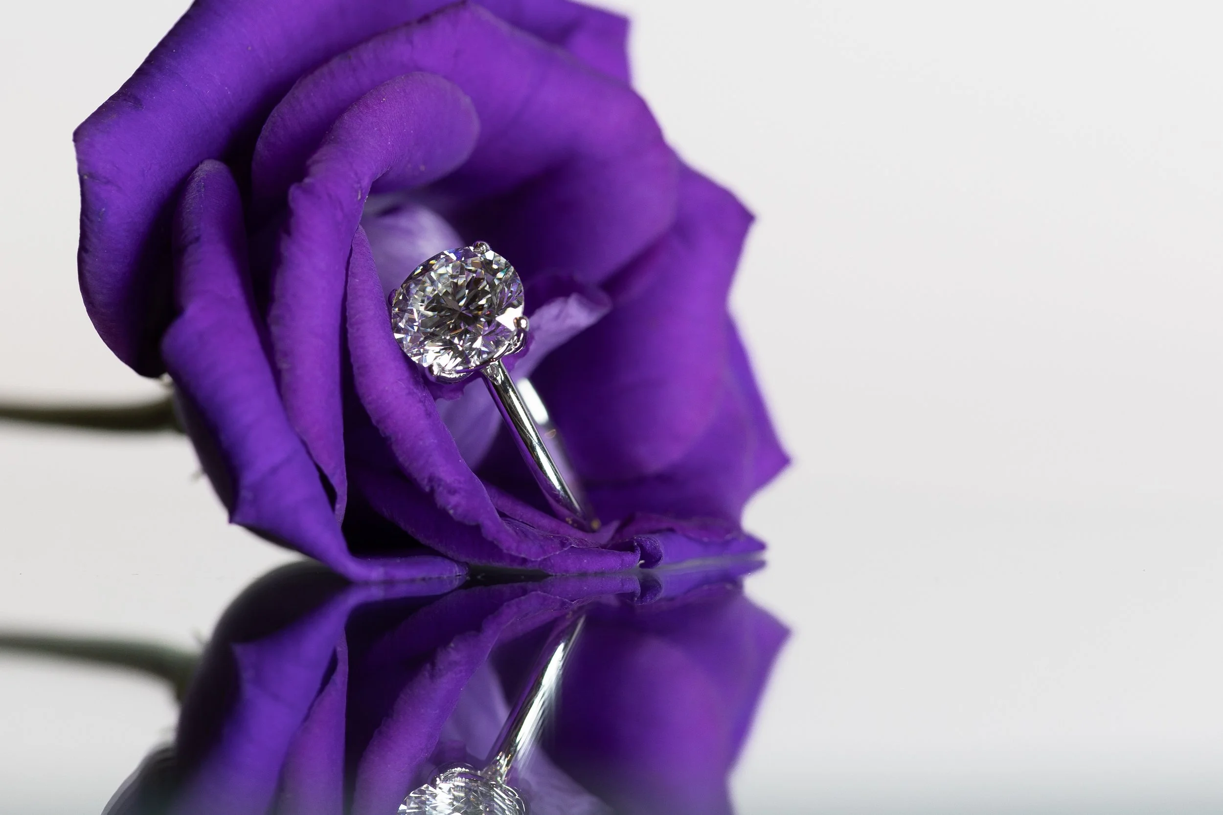 Een diamanten ring hangt aan een paarse roos met reflectie op een glad oppervlak.