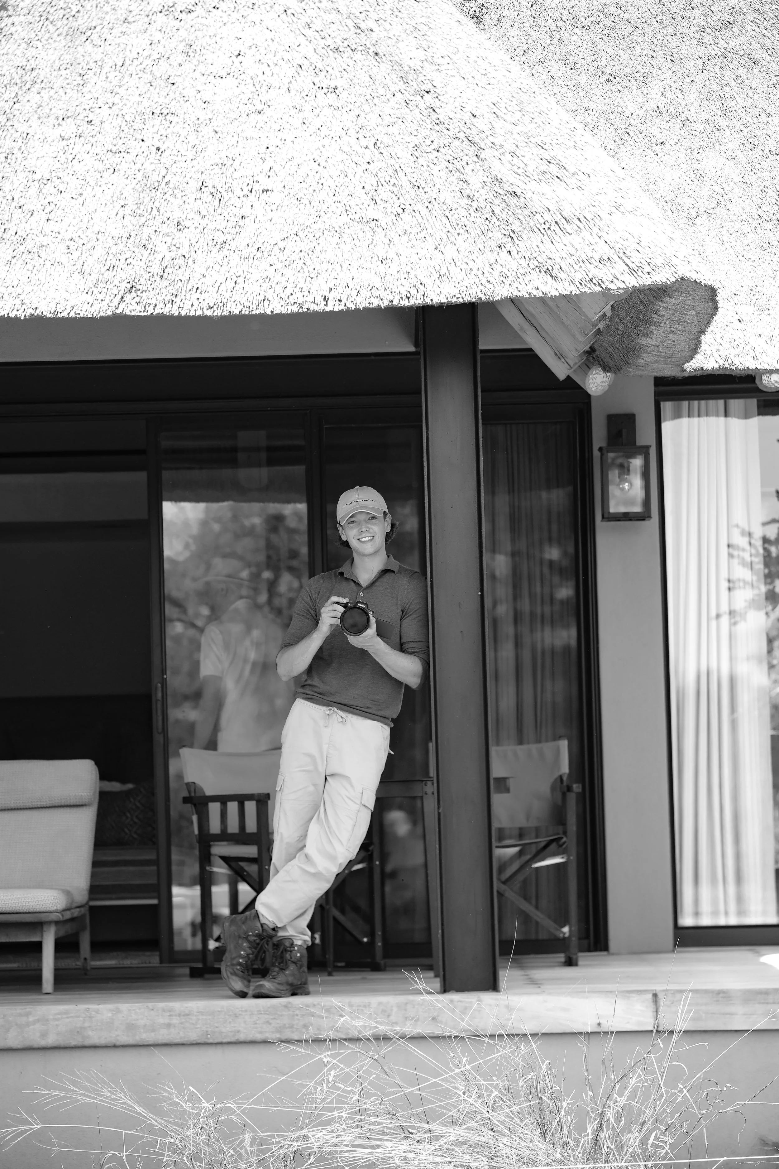 Persoon met camera die op een terras staat voor een woonhuis met grote ramen en een rieten parasol, zwart-wit foto.