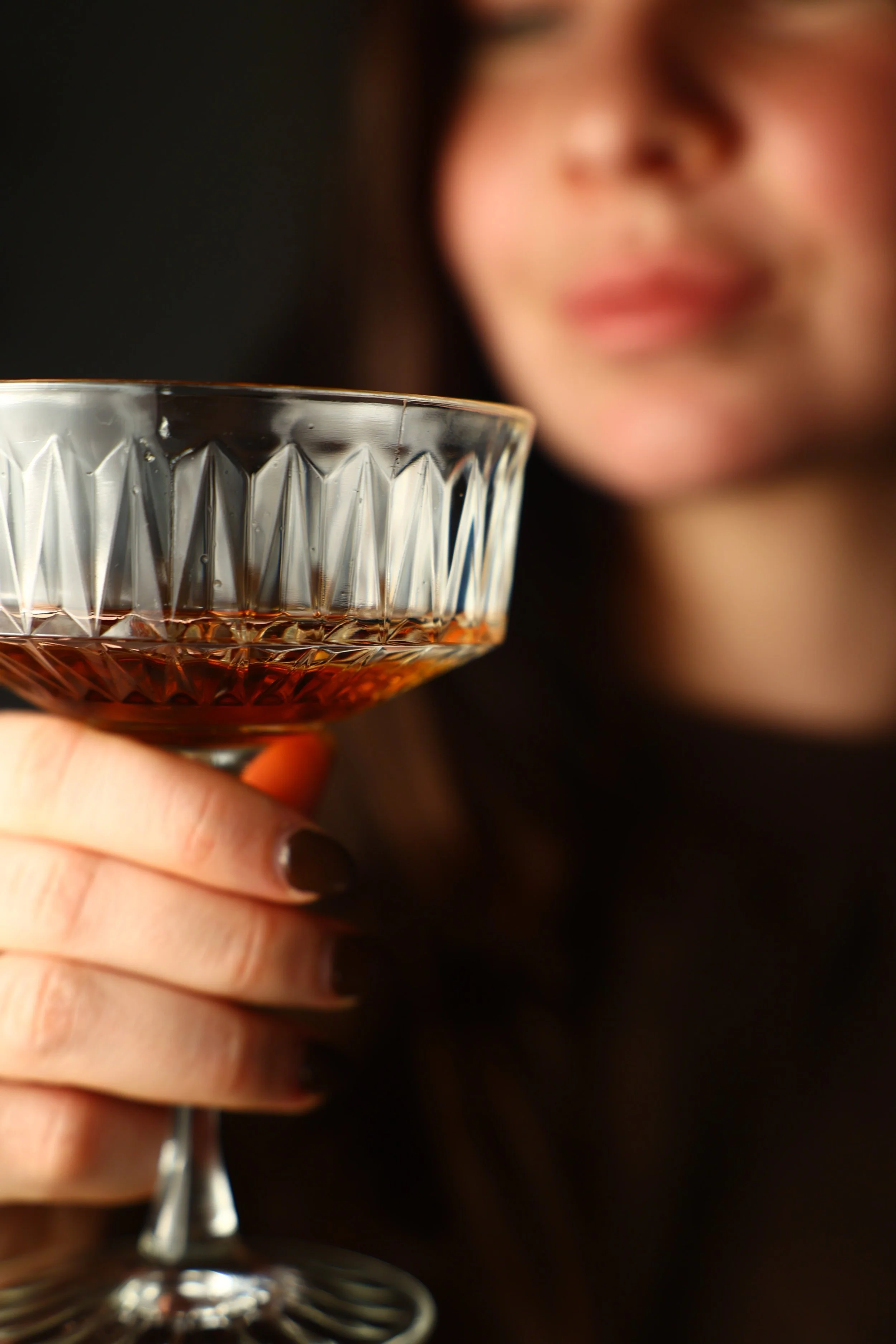 Een vrouw houdt een glas met een drank, waarschijnlijk heelal of sherry, in de hand, met een wazige glimlach op haar gezicht.