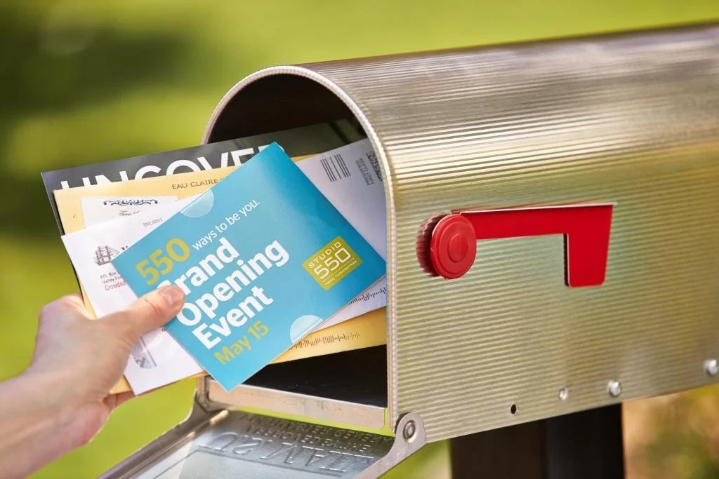 A person's hand placing a flyer into a mailbox. The flyer announces a grand opening event on May 15 with 550 ways to be you.