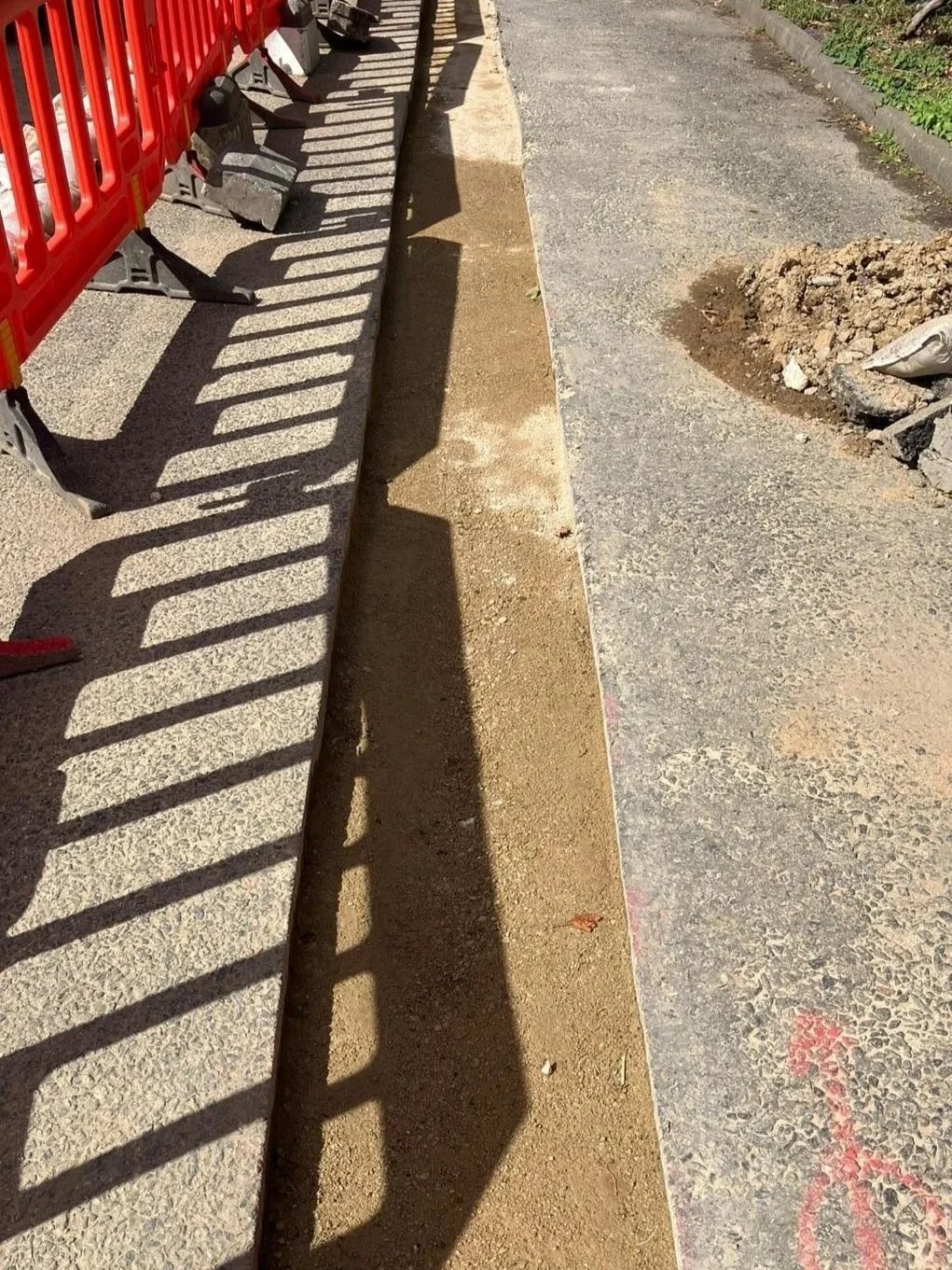 Construction site showing a new sidewalk being built, with a red safety barrier on the left and a dirt and gravel area on the right.