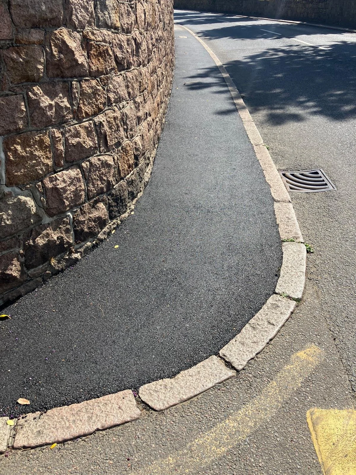 A sidewalk with a stone border along a curved street, a brick wall on the left, and a drainage grate on the right. Shadows of trees are cast on the street.
