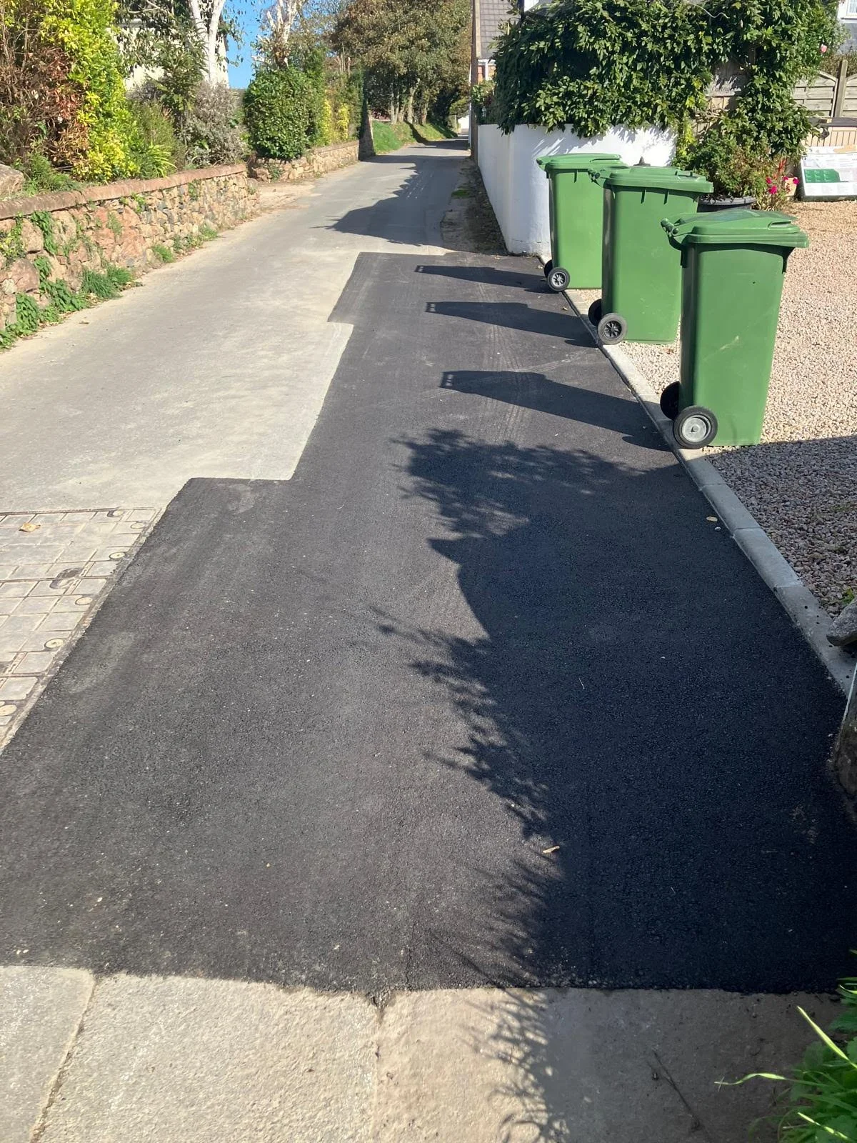 A residential street with green trash bins lined up along a white wall, trees and bushes on the right, and a sidewalk on the left with a section of new black asphalt pavement.