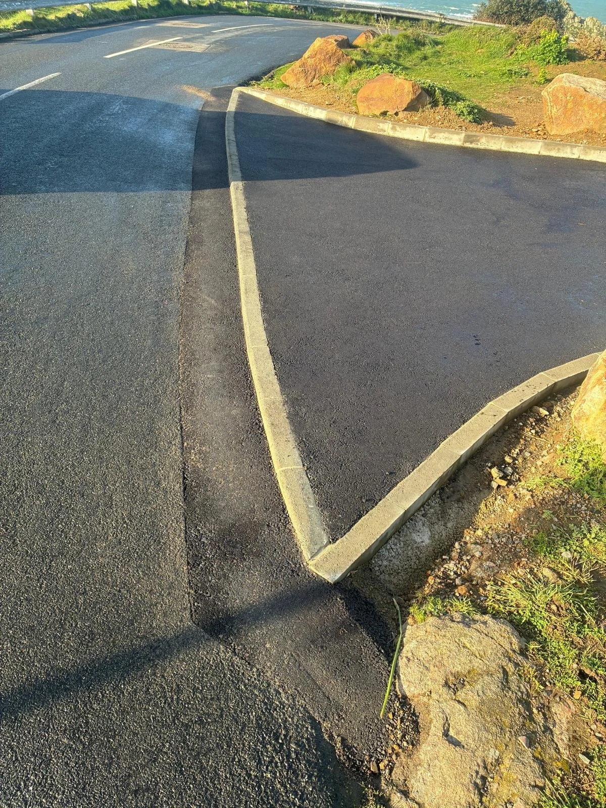 The image shows a newly paved asphalt parking lot with parking space lines and a curb with rocks and grass alongside it. There is greenery in the background and the sky is clear.
