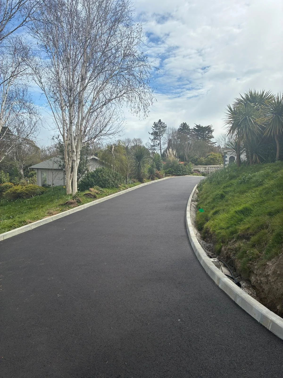 A freshly paved asphalt road winding through a suburban neighborhood with landscaped greenery and trees on either side, under a partly cloudy sky.
