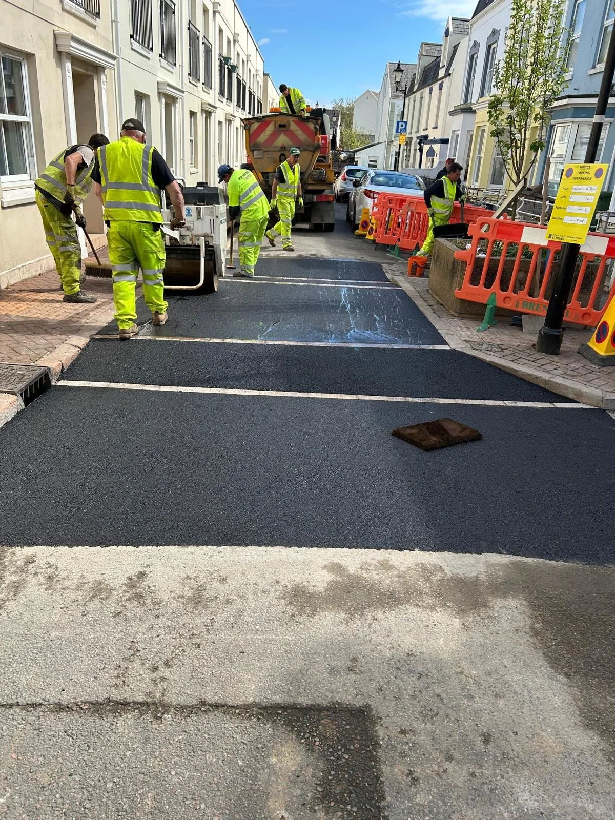 Construction workers in high visibility clothing are paving a new crosswalk on a city street. They are working with a paving machine and sandwiched orange safety barriers are set up along the sidewalk.