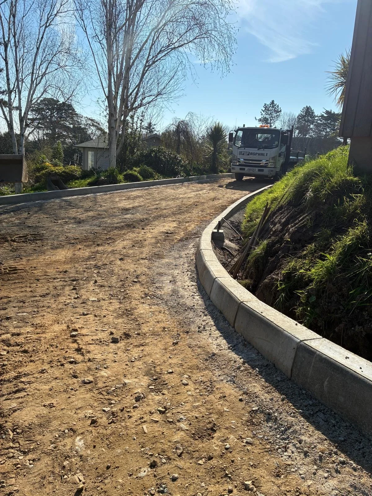 A dirt road under construction with a concrete curb on the right, a tree with bare branches, and a construction truck parked on the road. There are some houses and trees in the background and a clear blue sky.
