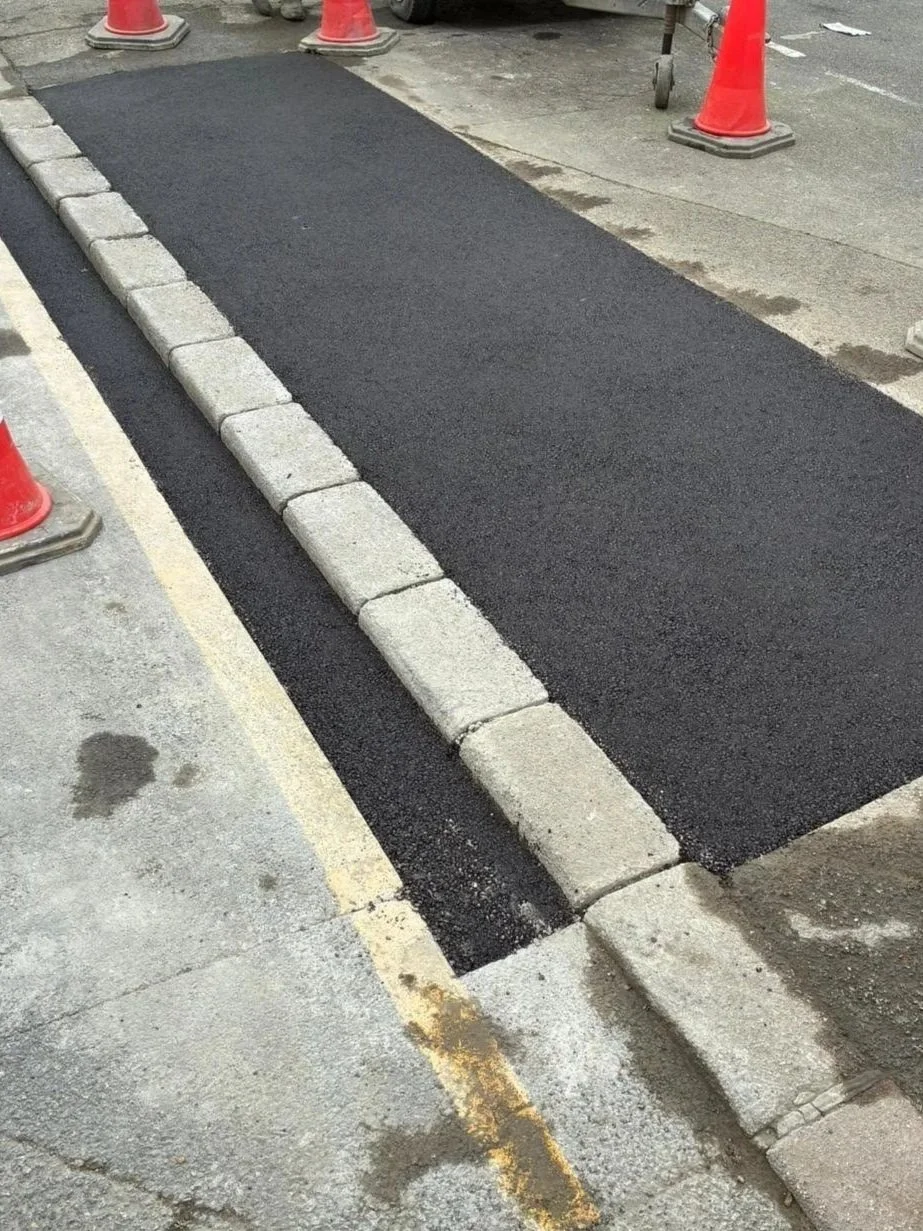 Freshly paved asphalt curb with concrete blocks on top, surrounded by orange traffic cones at a road construction site.