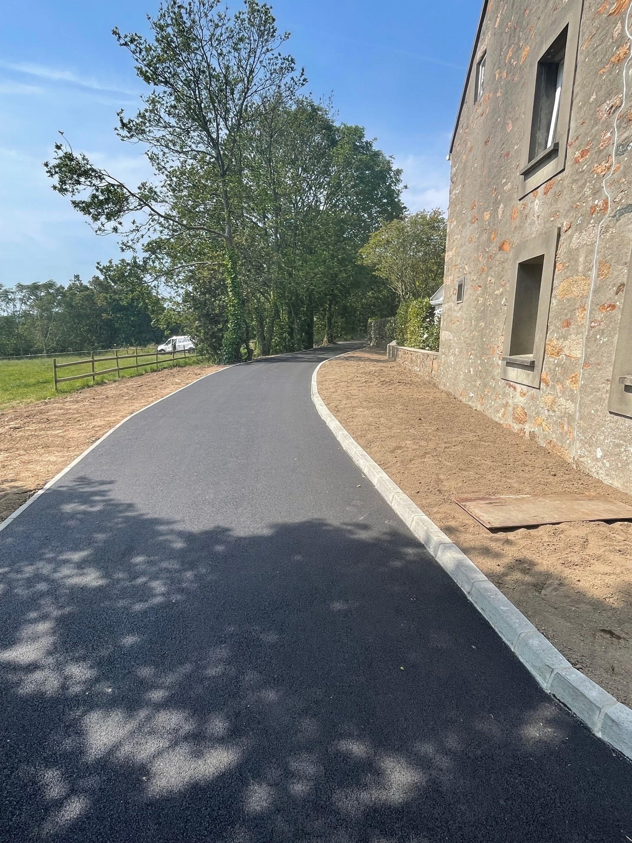 A newly paved asphalt road curves along the side of a stone building, with a grassy field and trees on the left, and the building's wall with windows on the right. A vehicle is visible in the background on the left side.