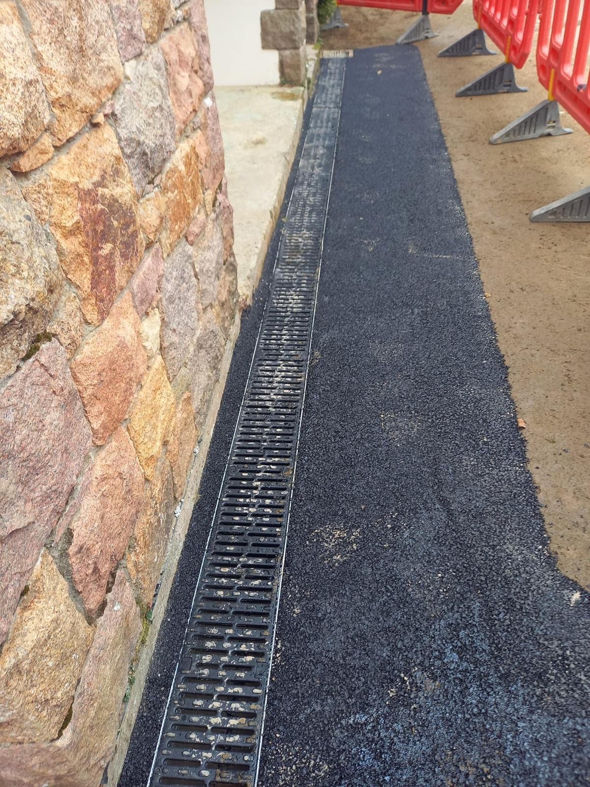 Newly paved black asphalt walkway with a metal drainage grate along the side, bordered by a stone wall on one side and orange construction fencing on the other, with dirt fencing visible in the background.