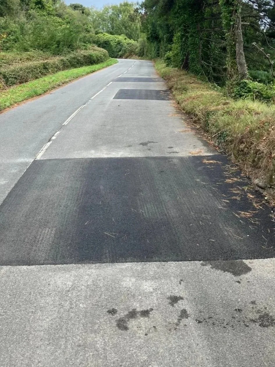 A rural road undergoing repairs with fresh asphalt patches placed on the road surface, flanked by greenery and trees on both sides.