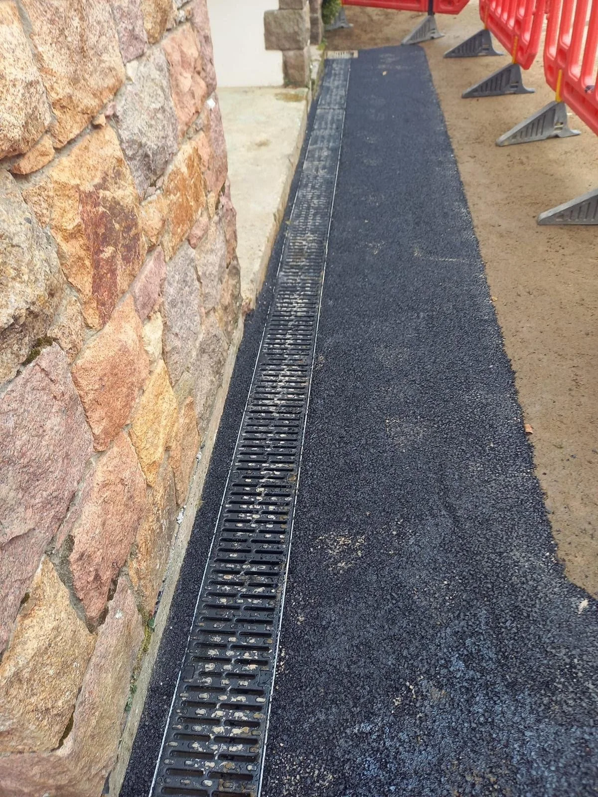 Close-up of a newly paved black asphalt sidewalk with a metal drainage grate along the edge, adjacent to a stone brick wall, and orange plastic safety barriers to the right.