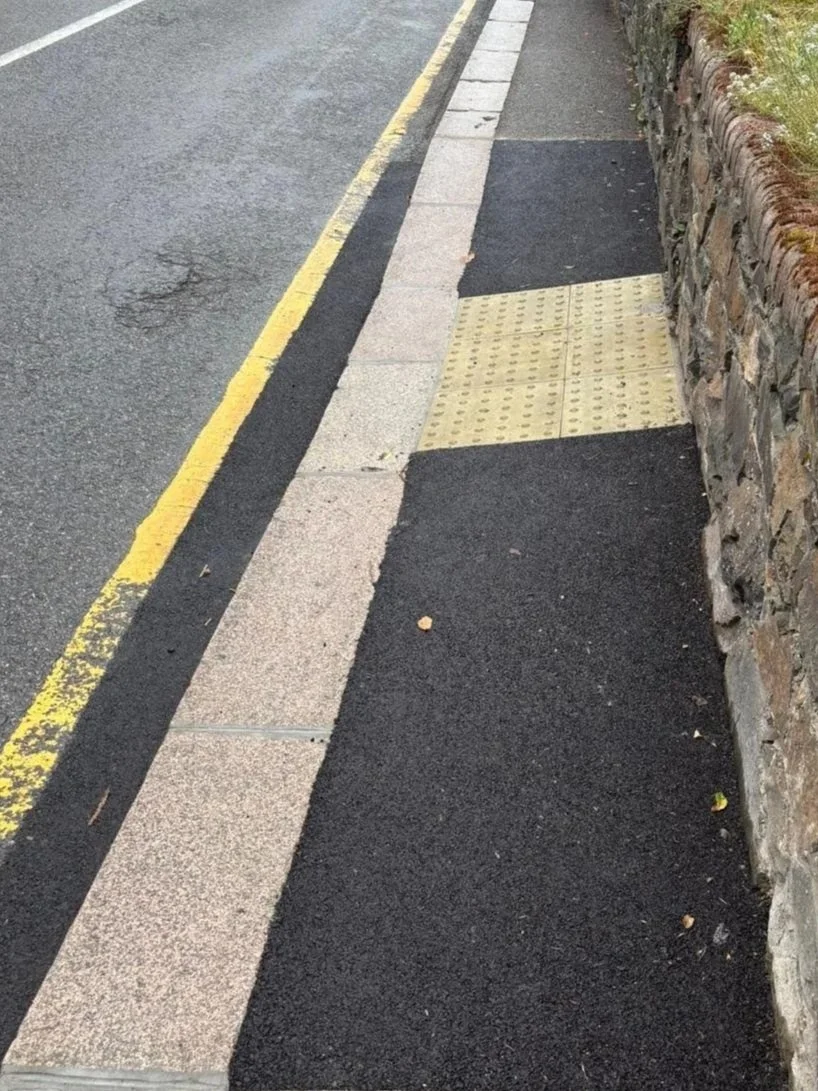 Close-up of a sidewalk with tactile paving tiles and a stone wall on the right, next to a street with a yellow line.