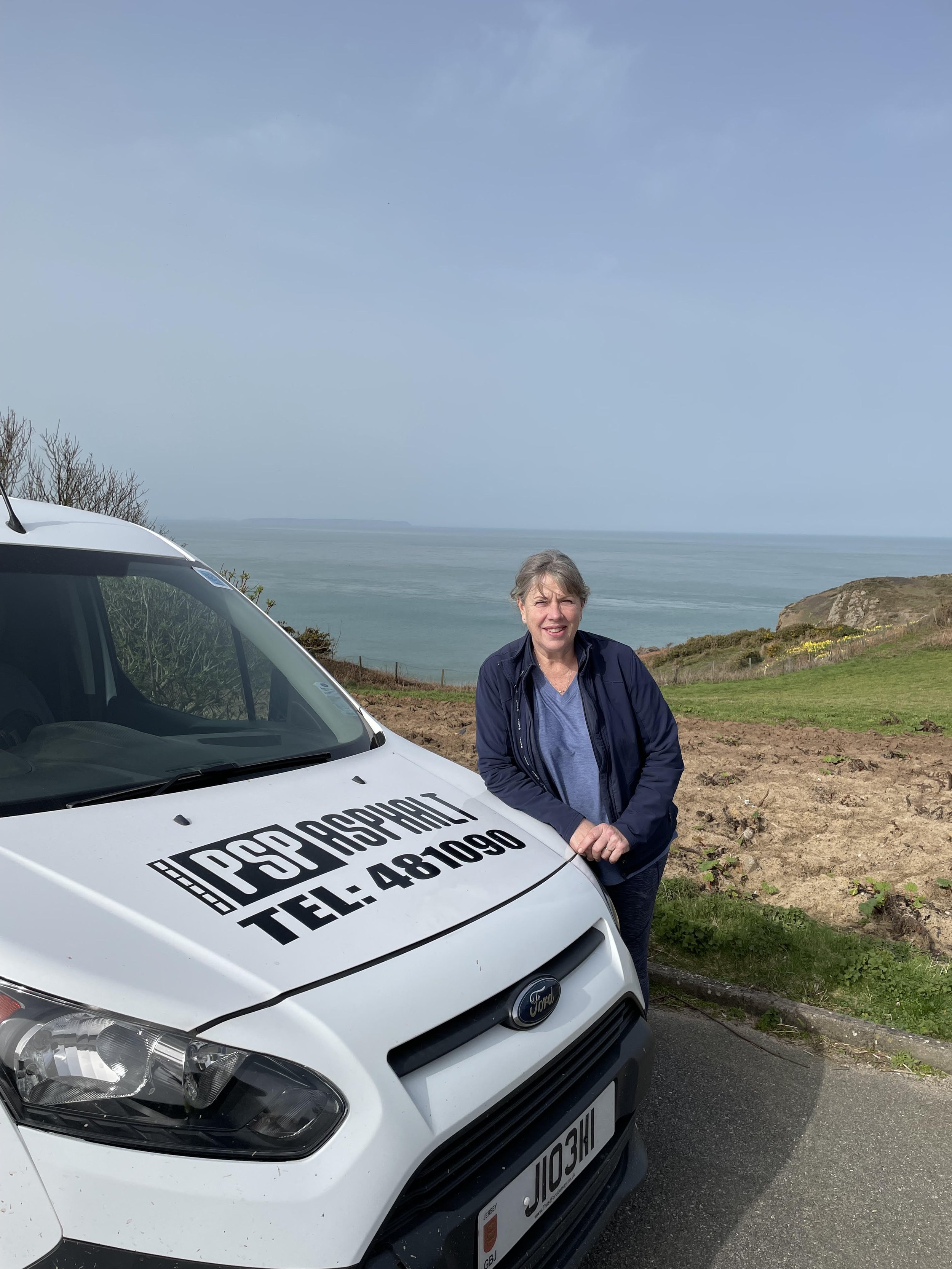 A woman standing beside a white Ford vehicle with a sign on the hood that reads PSP Asphalt with a coastal landscape and cloudy sky in the background.