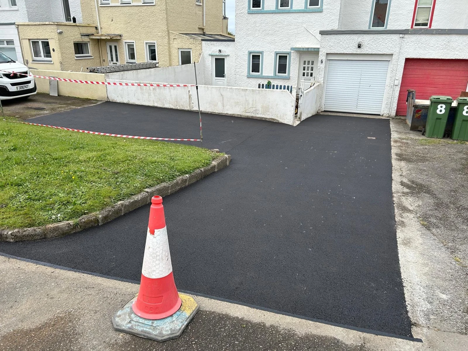 Freshly paved asphalt driveway with a safety cone and barrier tape, located in front of white and red garage doors, in a residential neighborhood with house facades and parked cars.