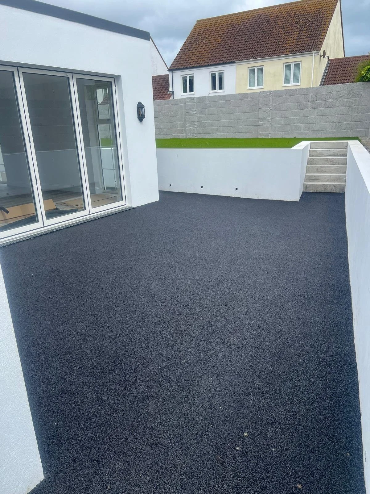 Newly constructed balcony or patio area with black rubberized flooring, white walls, and glass sliding doors leading into a house. In the background, a neighboring house with a red tile roof and gray concrete block wall is visible.