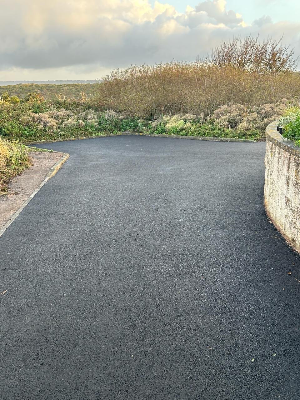 A newly paved black asphalt driveway curves to the right, bordered by a stone wall on the right and a garden with bushes and plants on the left, with a cloudy sky in the background.