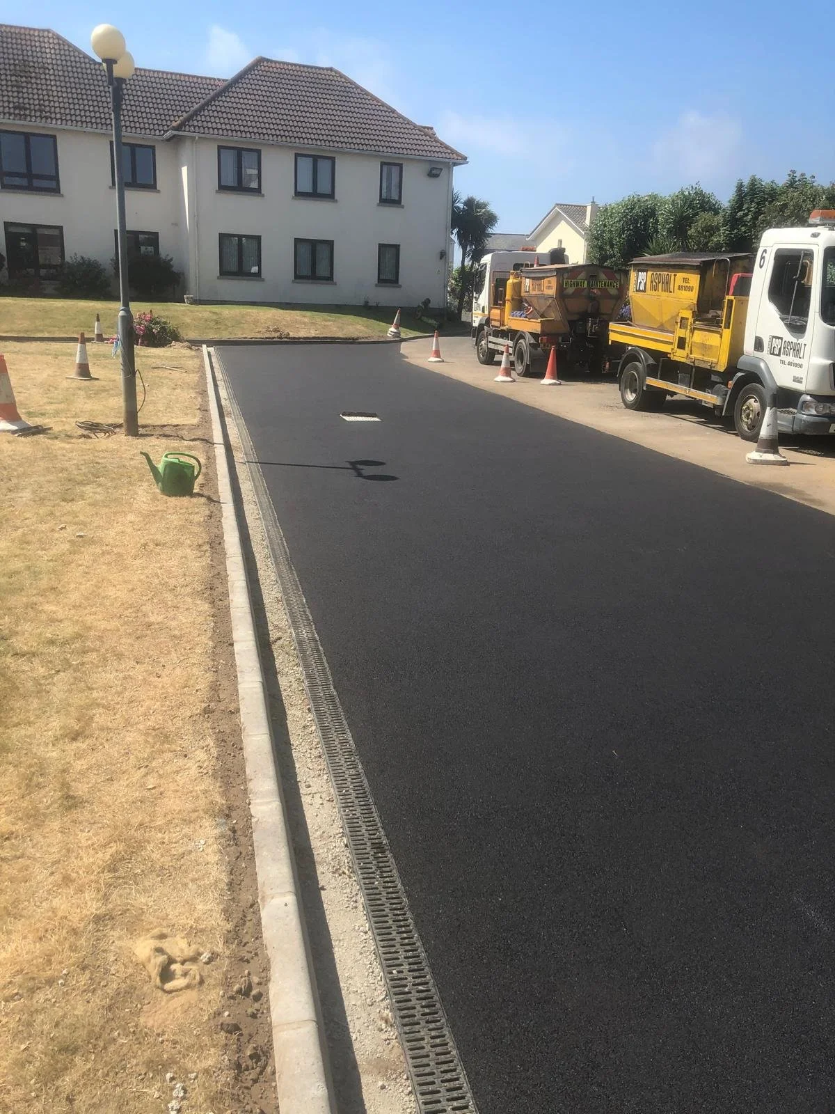 Newly paved black asphalt road in a residential area with construction vehicles and orange cones on the side. A watering can is placed on the grass, and there is a multi-story house in the background.