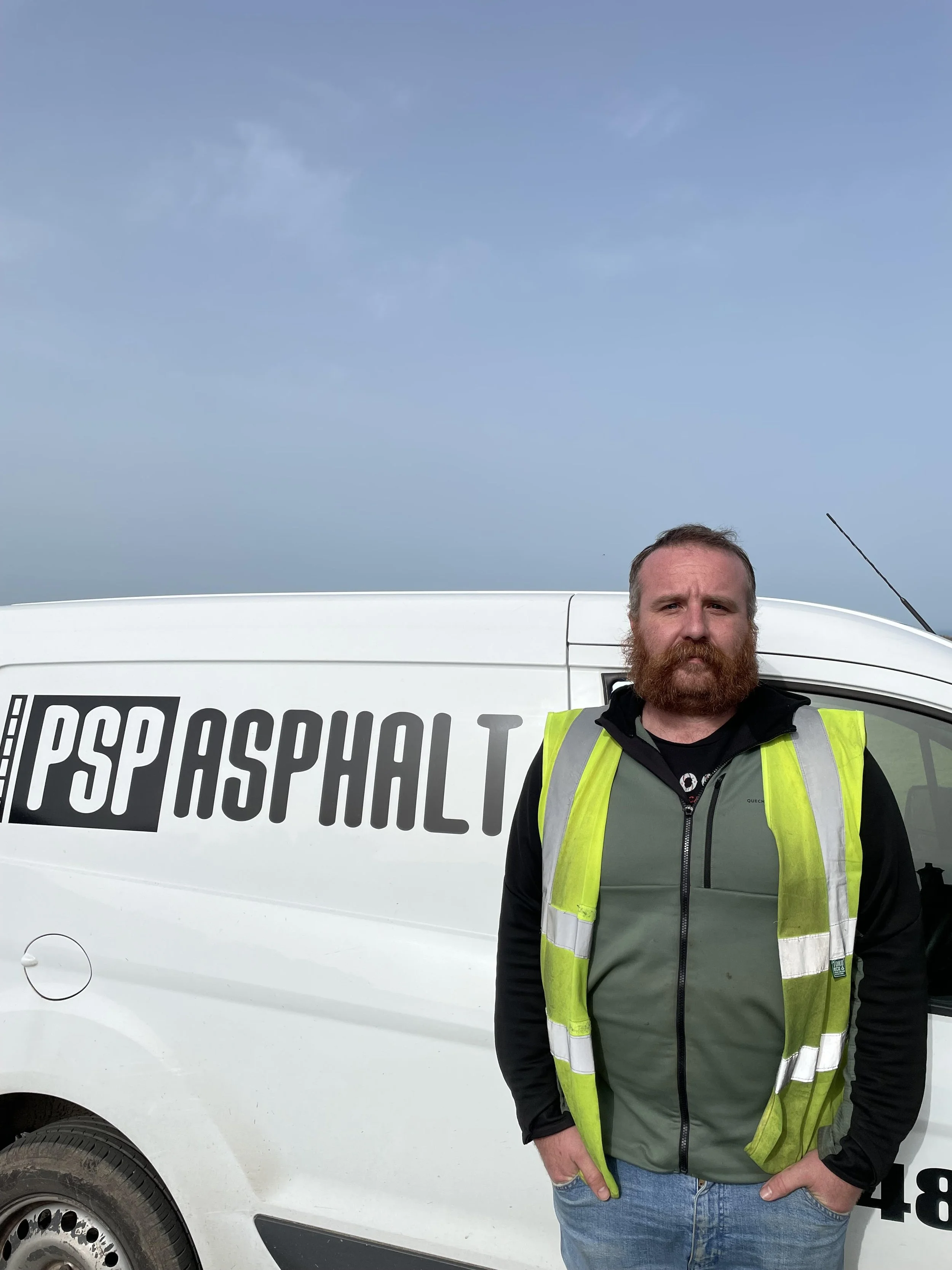 A man with a beard and casual clothing wearing a yellow safety vest stands in front of a white vehicle with partially visible text. The background shows a clear blue sky.