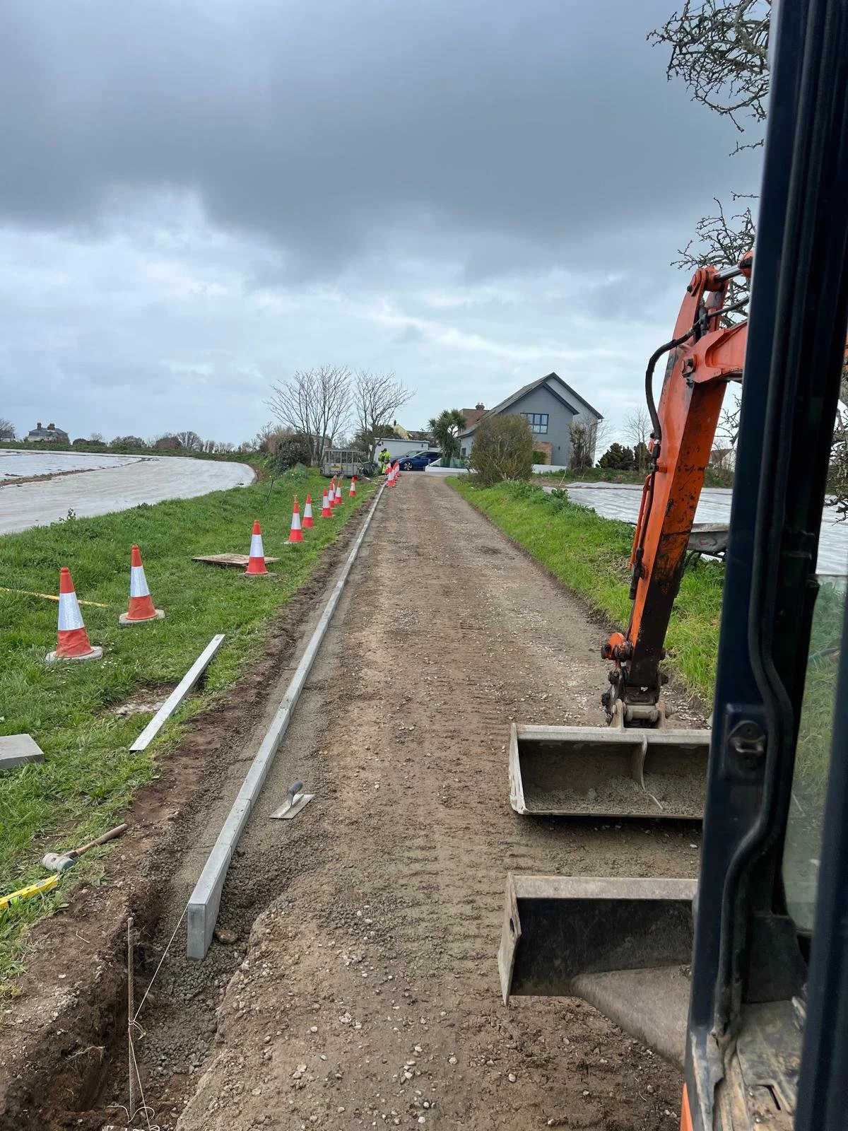 A construction site on a dirt pathway, with orange traffic cones on the left side, construction equipment on the right, and a house in the background under a cloudy sky.