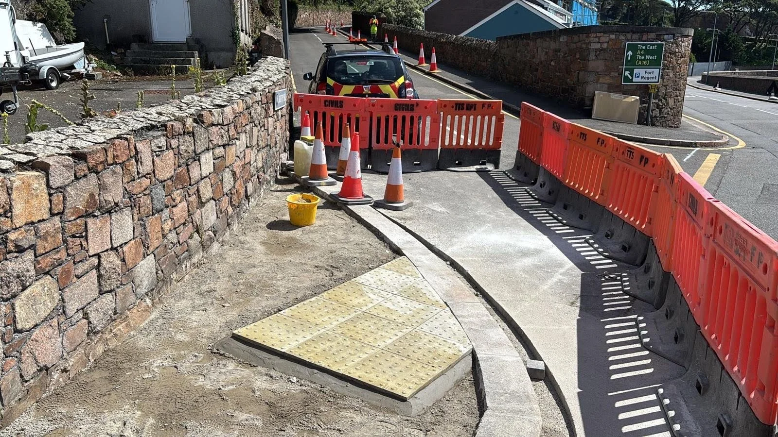 Street construction site blocked by orange safety barriers and traffic cones, with a parked vehicle behind the barriers on a narrow street, and a green directional road sign in the background.