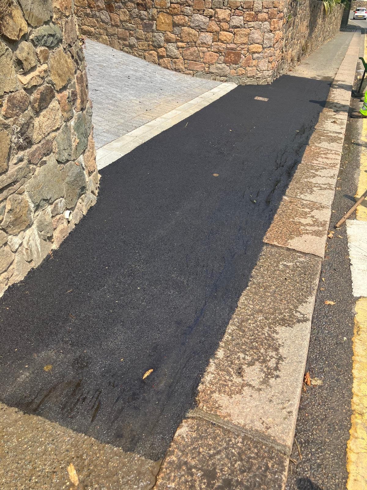 Freshly paved black asphalt sidewalk next to a stone wall and curb, with a section of gray brick pavement nearby.