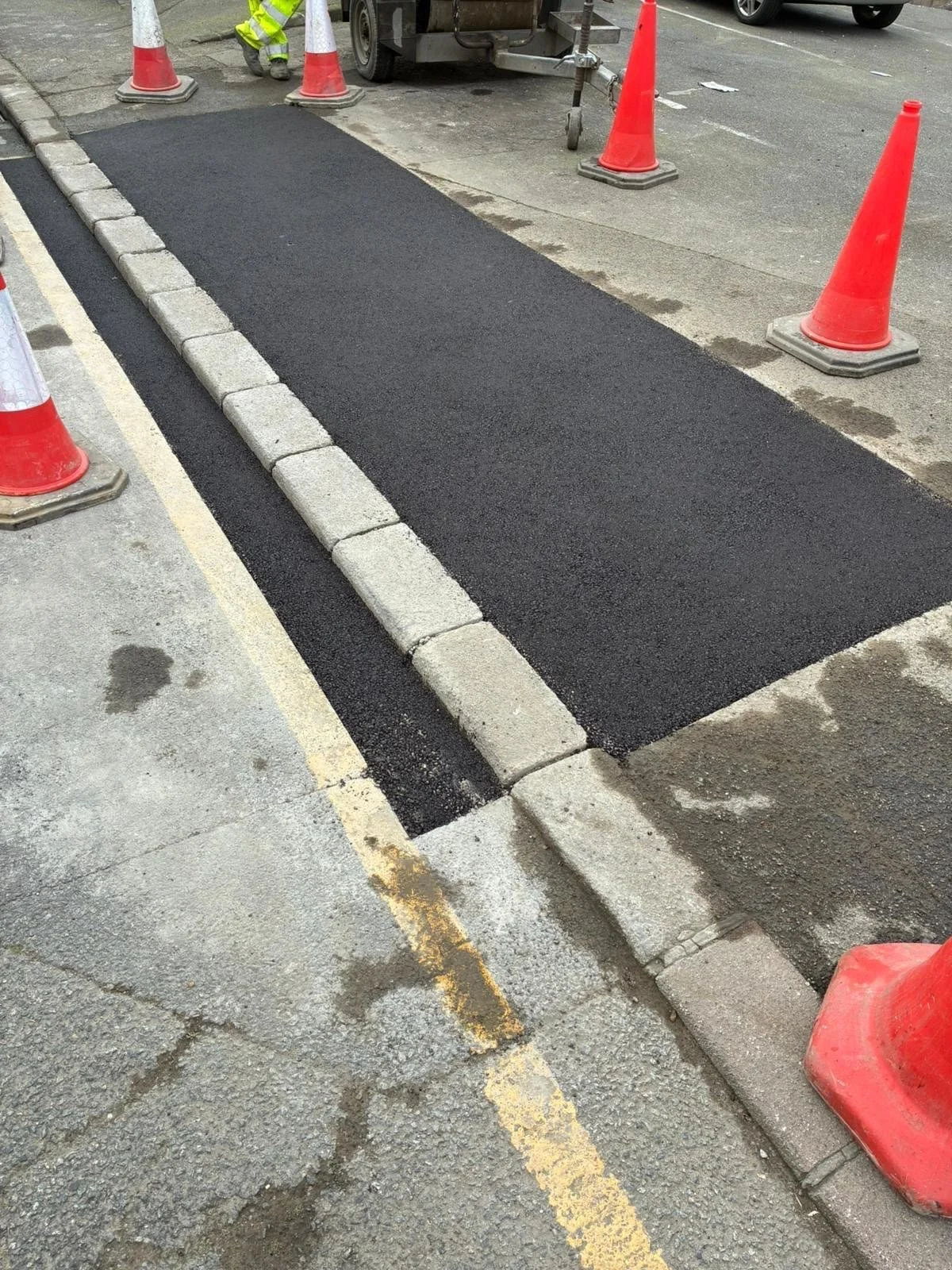 Freshly paved asphalt section on a street with orange traffic cones around it, and a worker in yellow safety gear visible at the top of the image, near construction equipment.