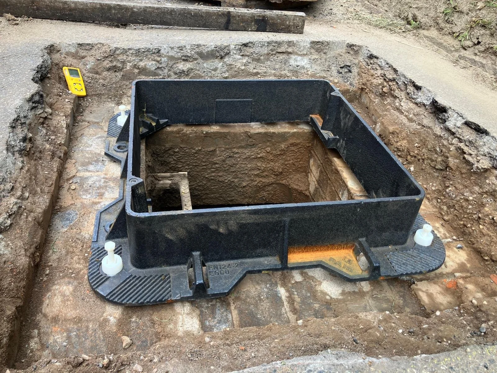 Excavation site with a black utility access box in the ground, some construction tools and a digital level nearby, dirt and concrete surrounding the area.