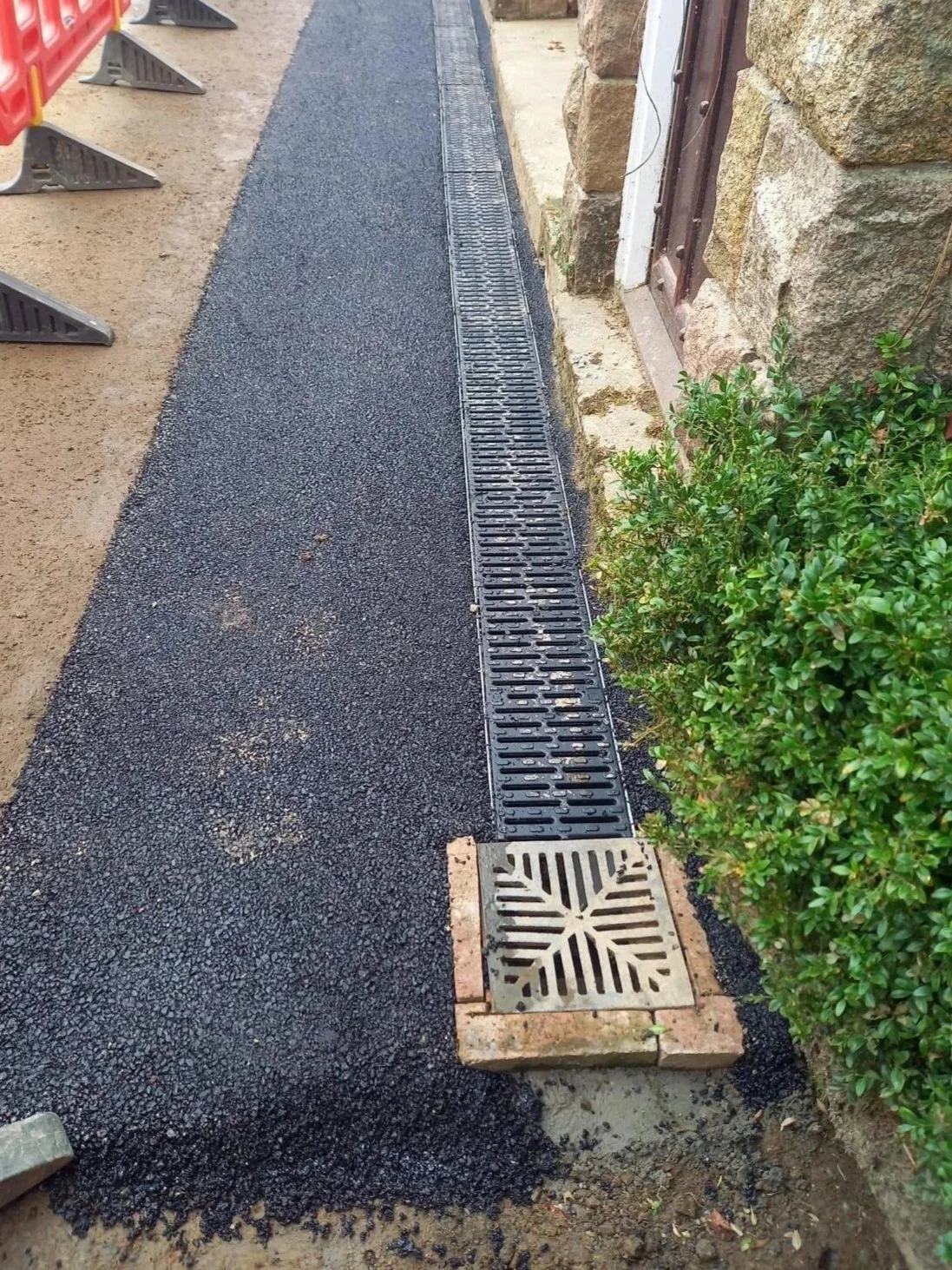 Freshly paved asphalt sidewalk with a drainage grate and a decorative drain cover, bordered by a stone wall and green shrubbery.