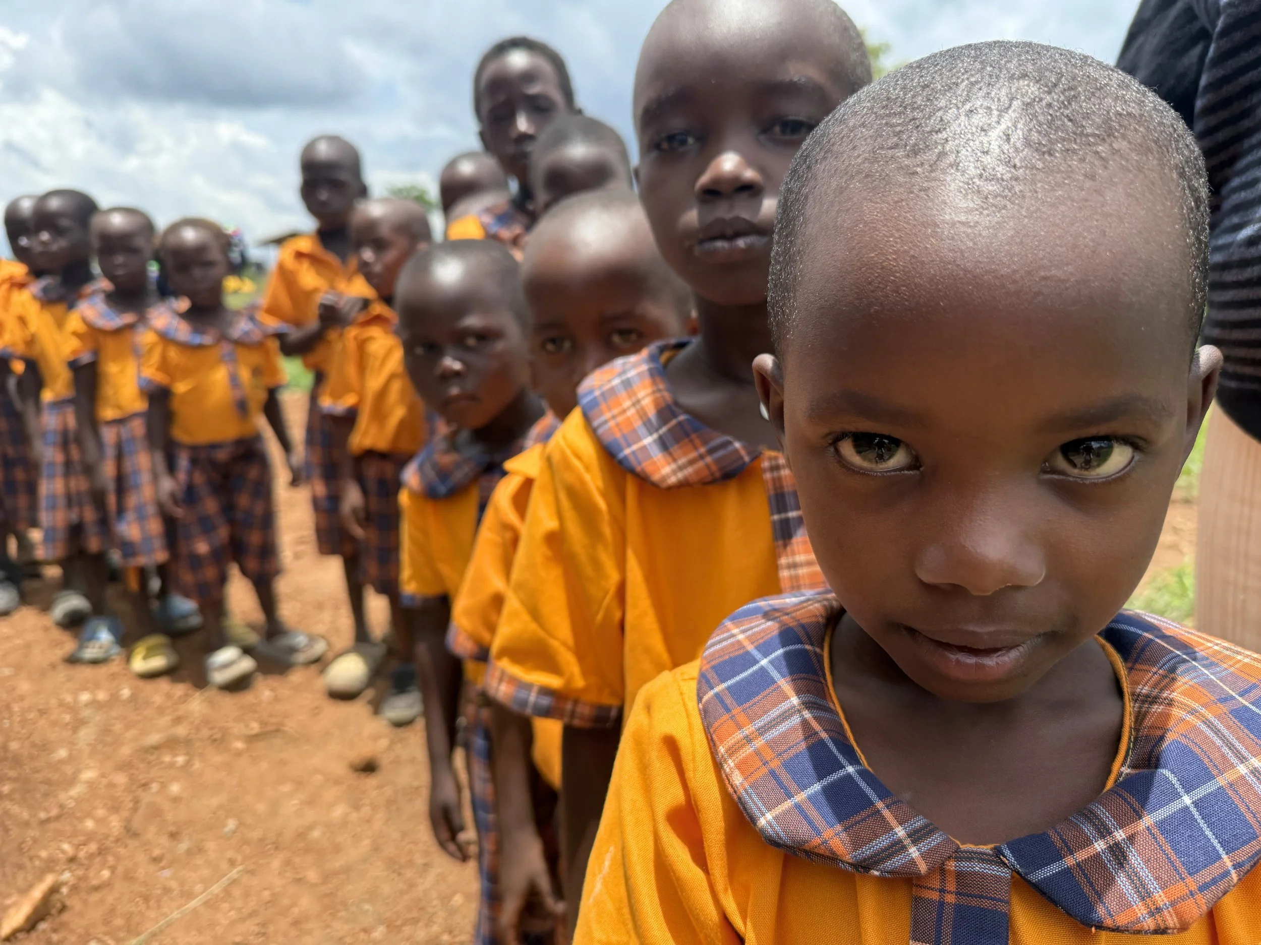 Bambini africani in uniforme scolastica con linee ordinate su una riga, con uno sguardo diretto verso la fotocamera, in un ambiente all'aperto con cielo nuvoloso.