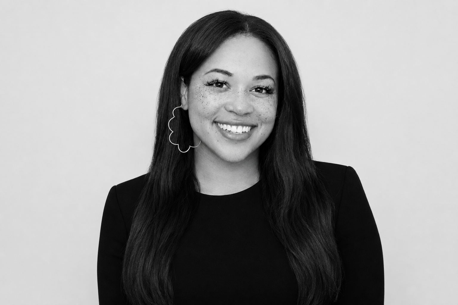 Black and white portrait of a woman with long dark hair, large hoop earrings, smiling, wearing a dark top, plain background.
