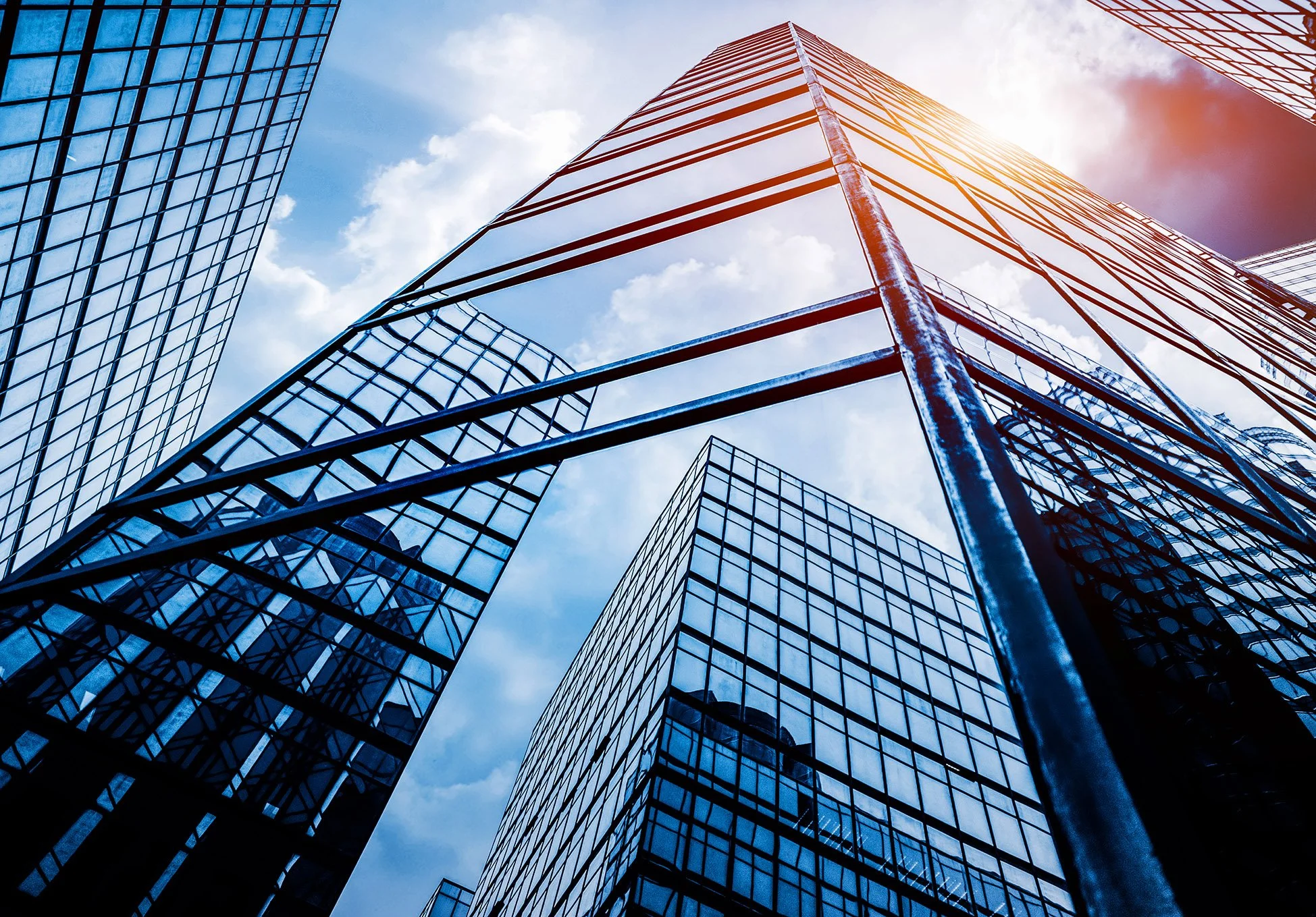 Low-angle view of modern glass skyscrapers against a blue sky with clouds and sunlight.
