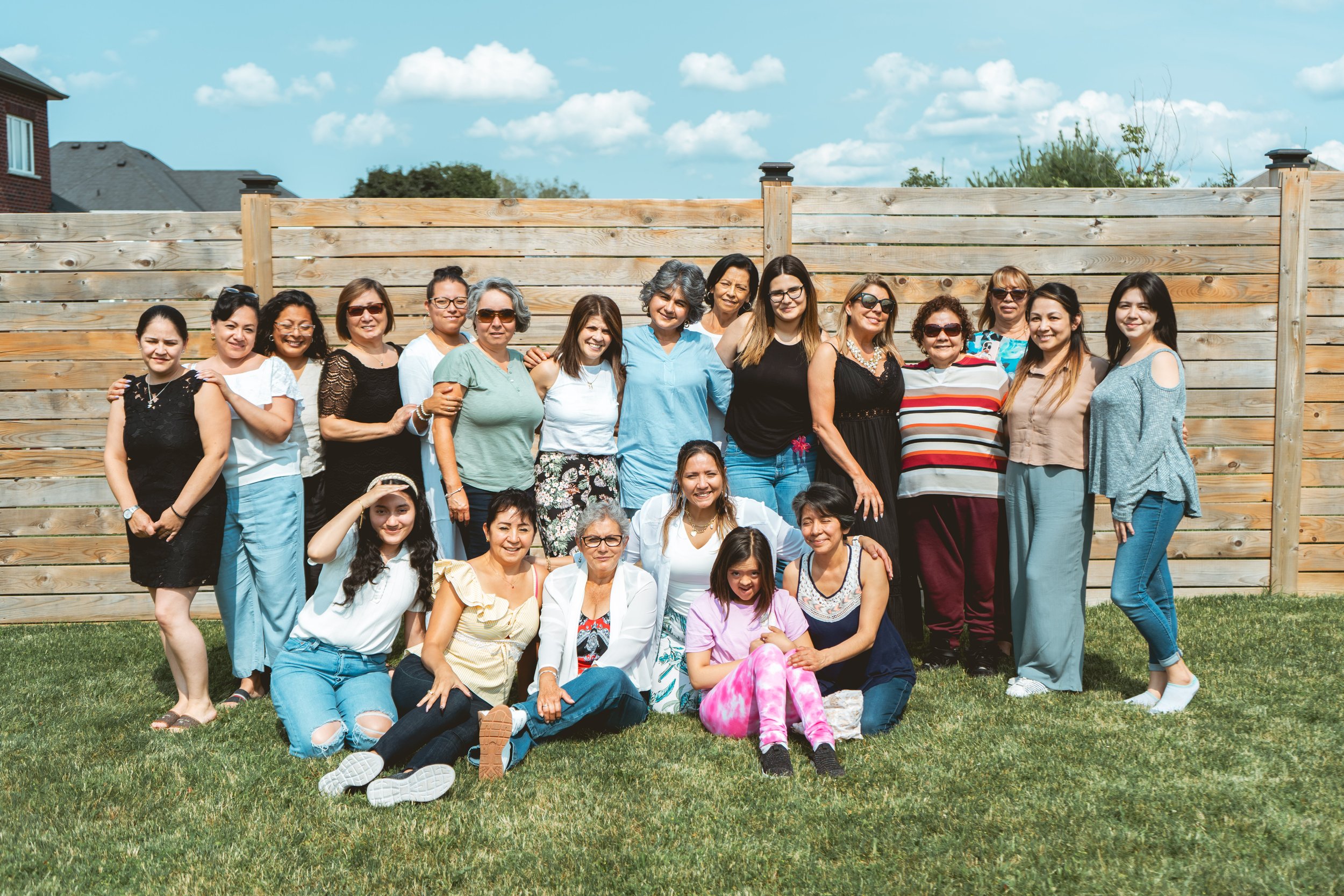A group of women posing in a garden, with some sitting on the grass and others standing; a wooden fence is in the background.