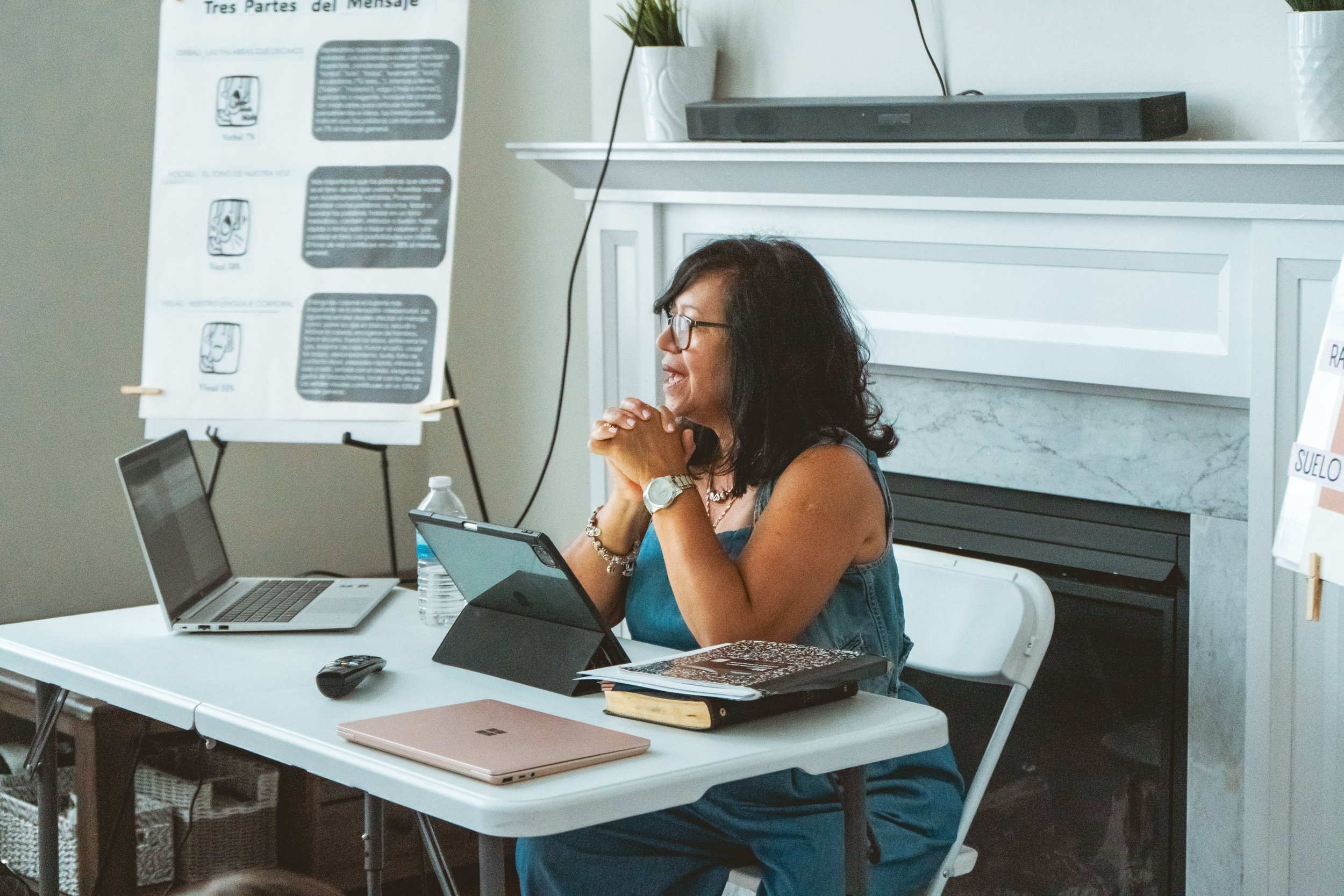 A woman sitting at a desk, speaking or teaching, with a laptop, water bottle, notebook, and tablet on the table. In the background, there is a poster with Spanish text and diagrams propped up on an easel.