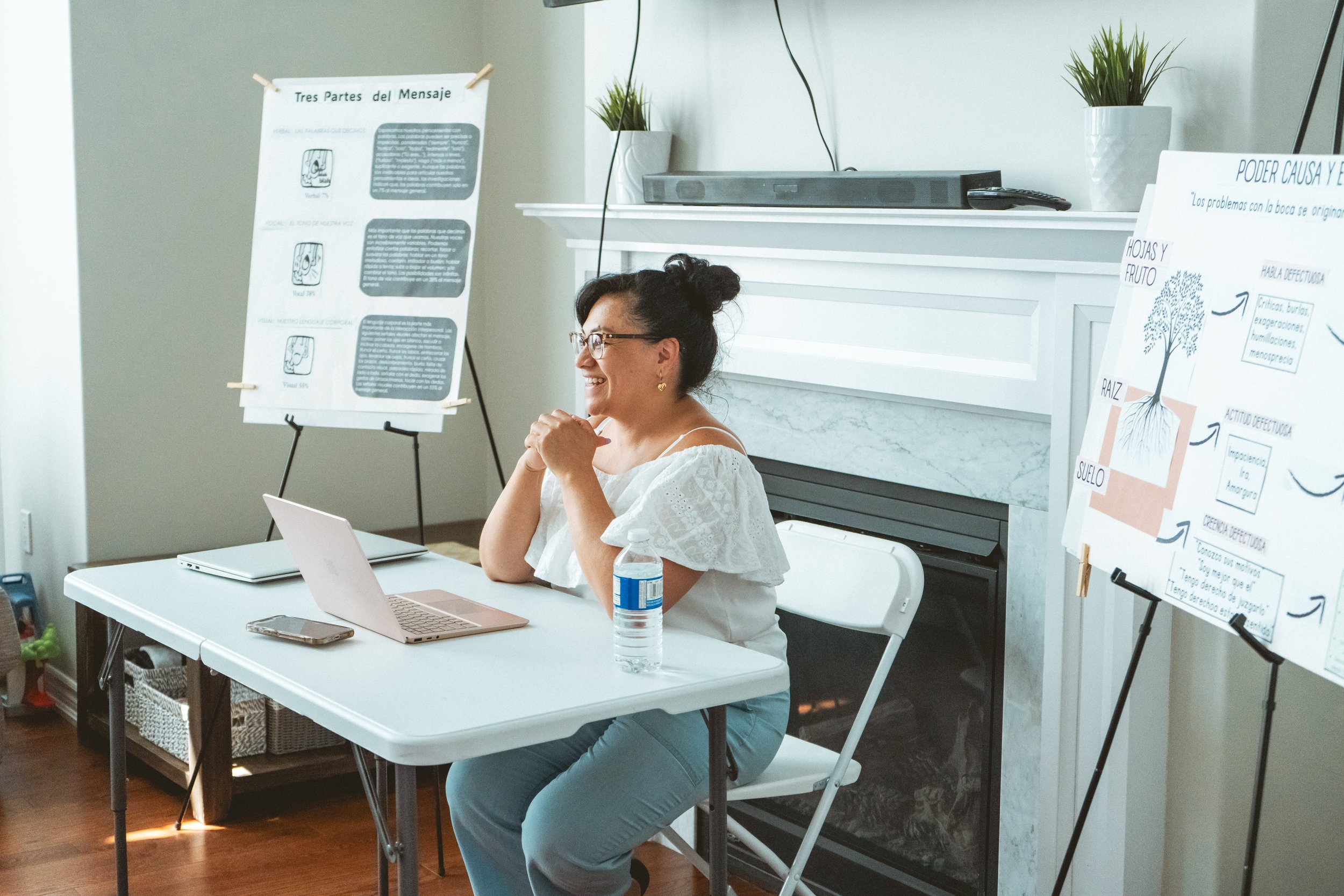 A woman sitting at a table with a laptop and a water bottle, conducting a presentation with charts on display boards in a home setting.